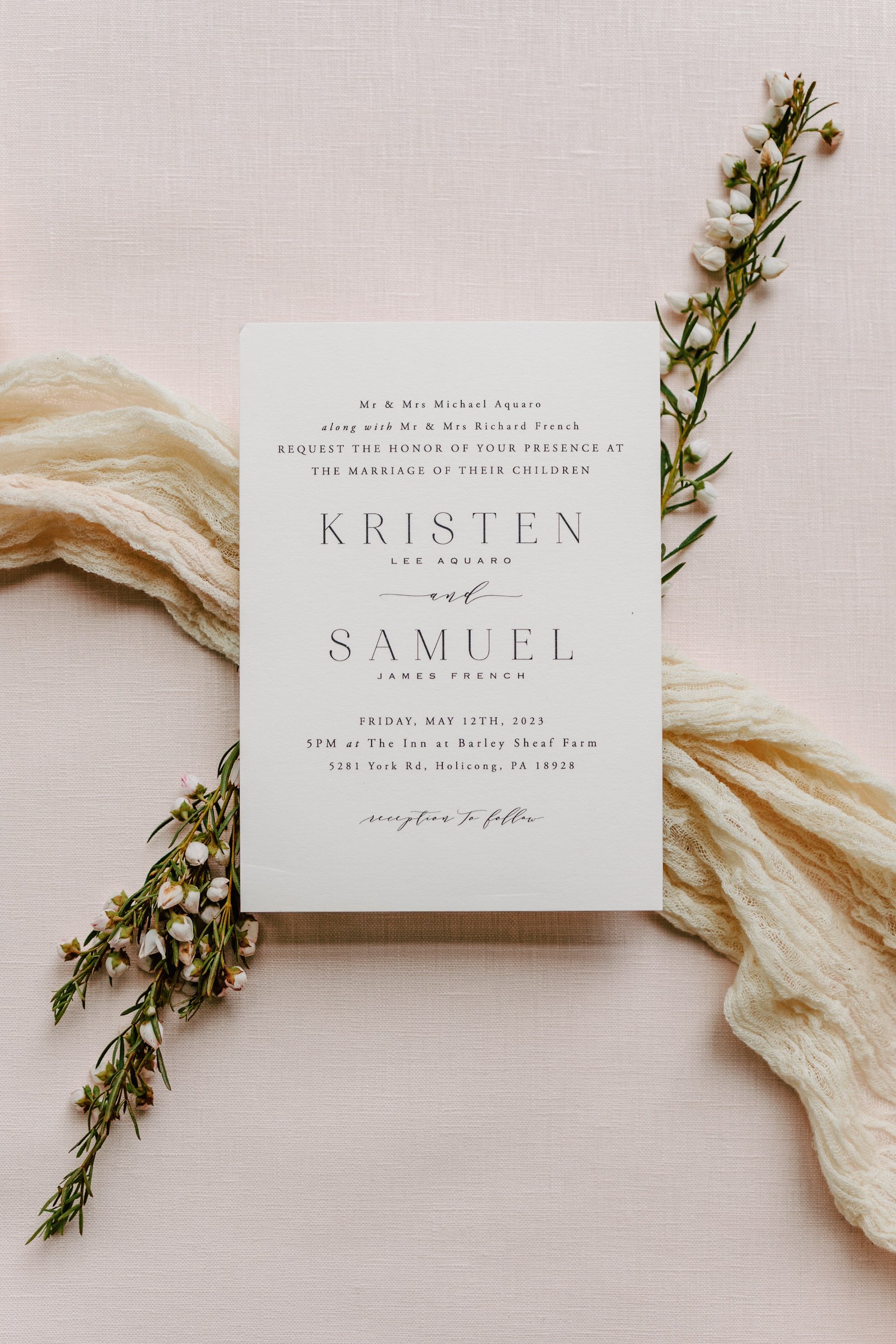 Wedding invitation card placed on a beige fabric surface, accompanied by a sprig of white flowers and a cream-colored fabric ribbon.