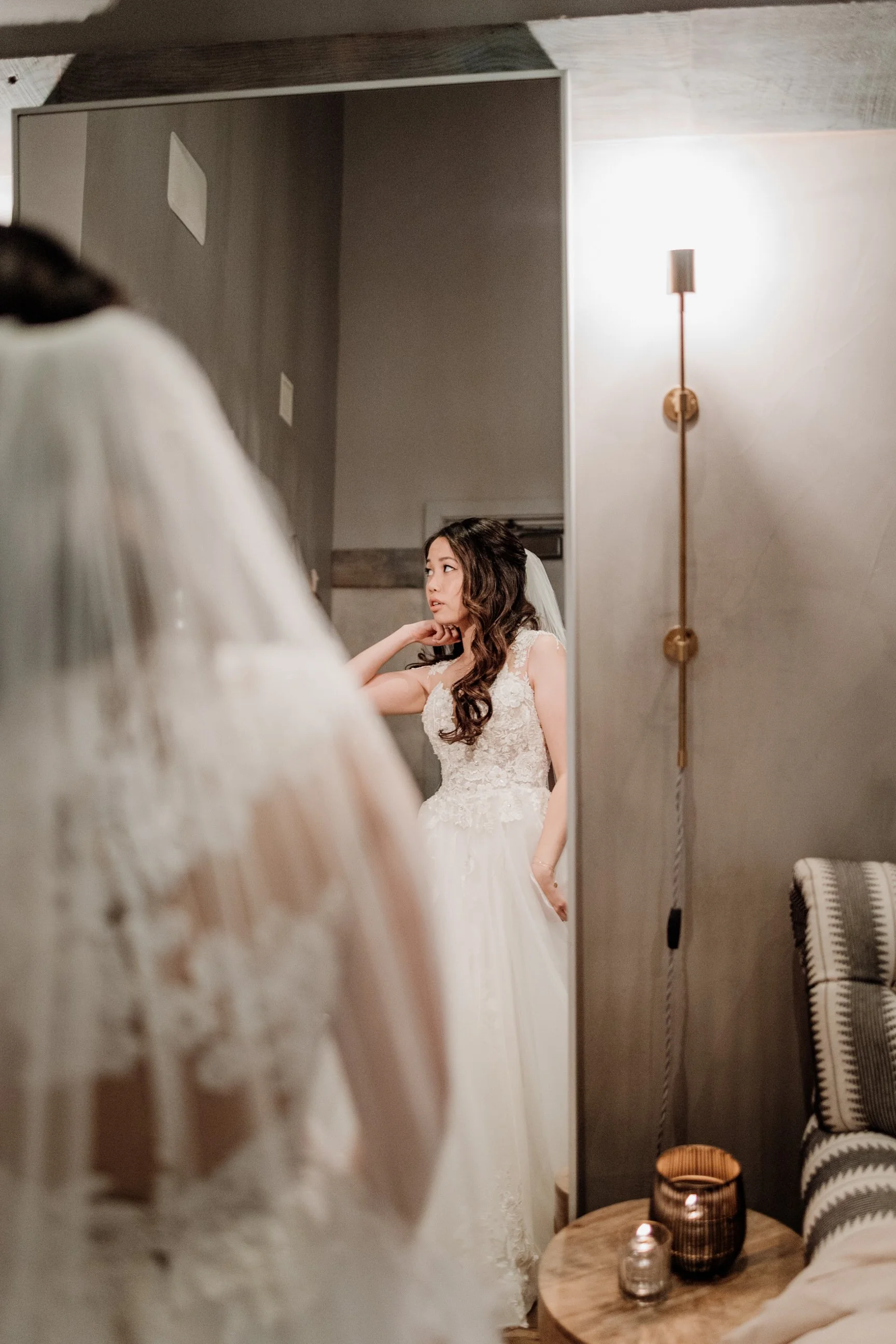 A bride in a white wedding dress looking at herself in a large mirror, with a reflection showing her sitting with her hand on her neck.