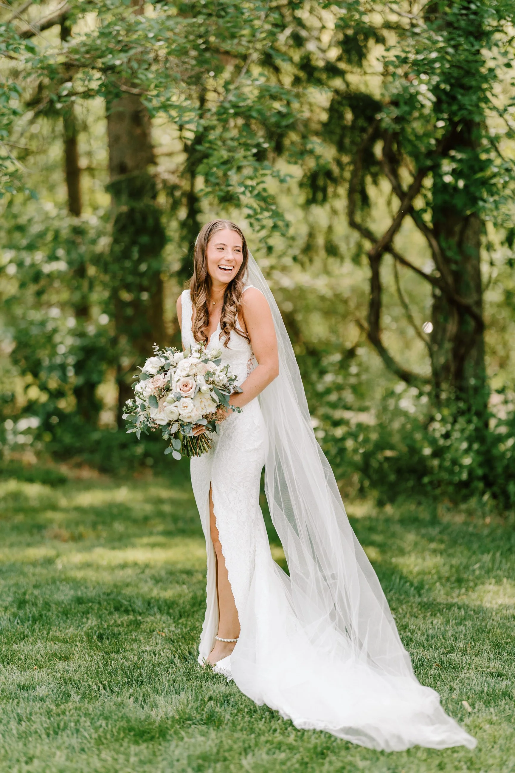 A joyful bride in a white lace wedding gown with a slit, holding a large bouquet of white and soft pink flowers, standing on green grass in an outdoor, wooded setting.