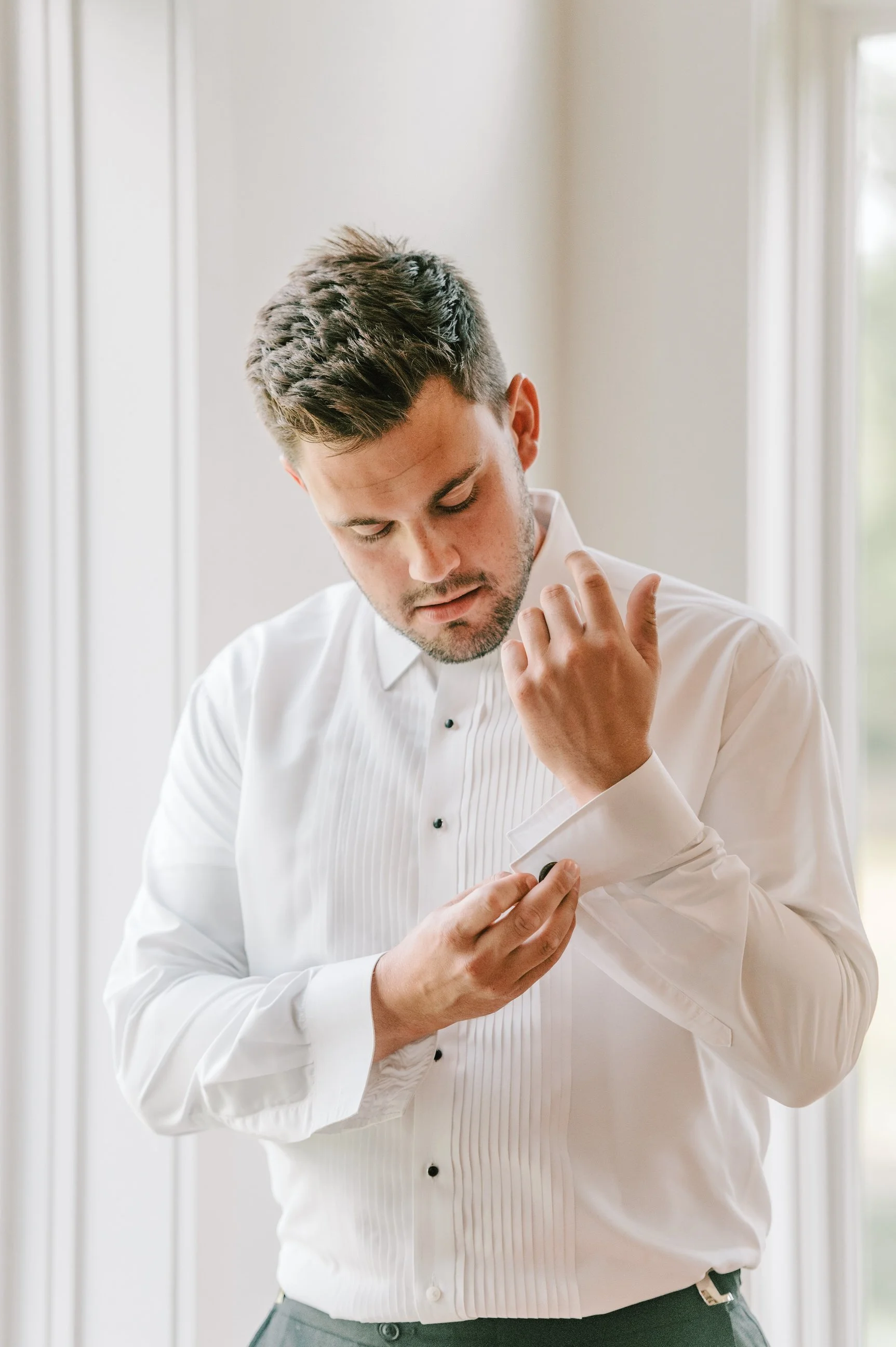 A man in a white dress shirt adjusting his cufflink, standing near a window.