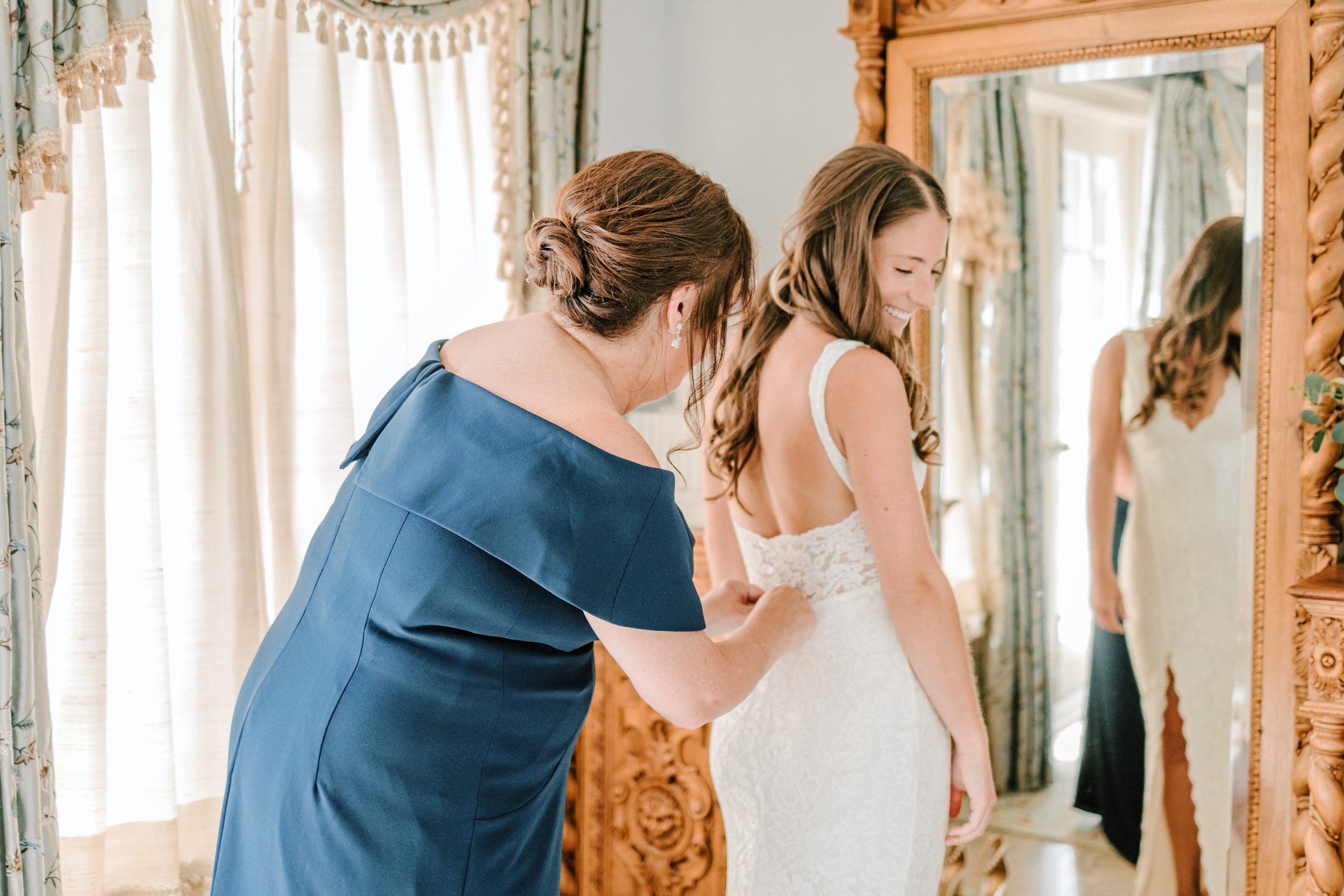 A bride in a white lace gown smiling as an older woman, possibly her mother, adjusts her dress in front of a mirror in a well-lit room with elegant curtains.