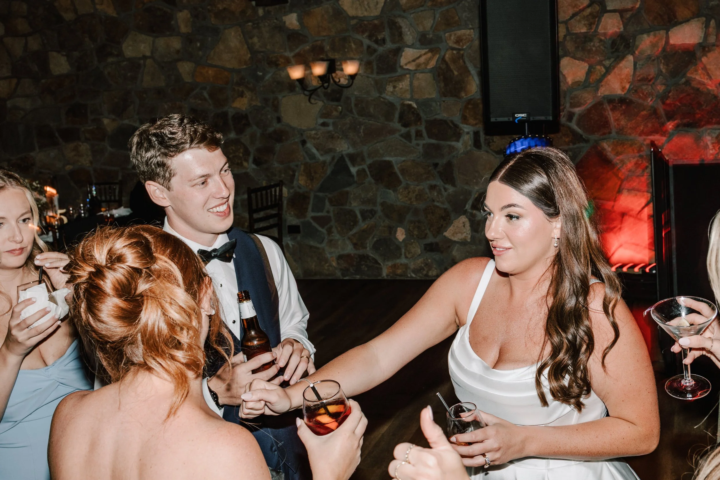 Group of people at a party, with women in dresses and a man in a tuxedo, enjoying drinks and socializing in a room with a stone wall and dim lighting.