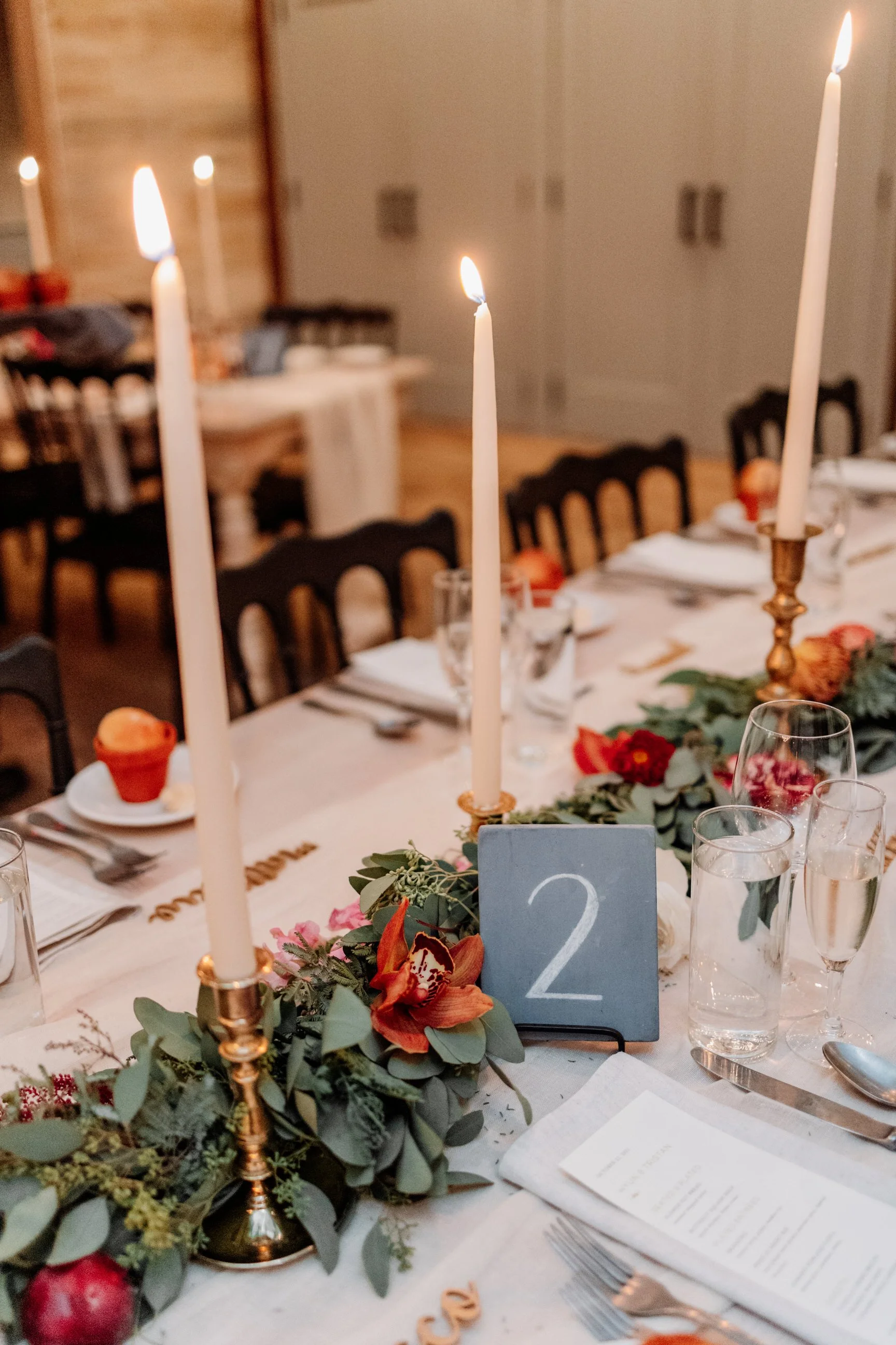 Elegant dining table decorated with greenery, orange flowers, tall white candles in brass holders, glassware, plates, utensils, and a table number sign with the number 2, set for a celebration or wedding.