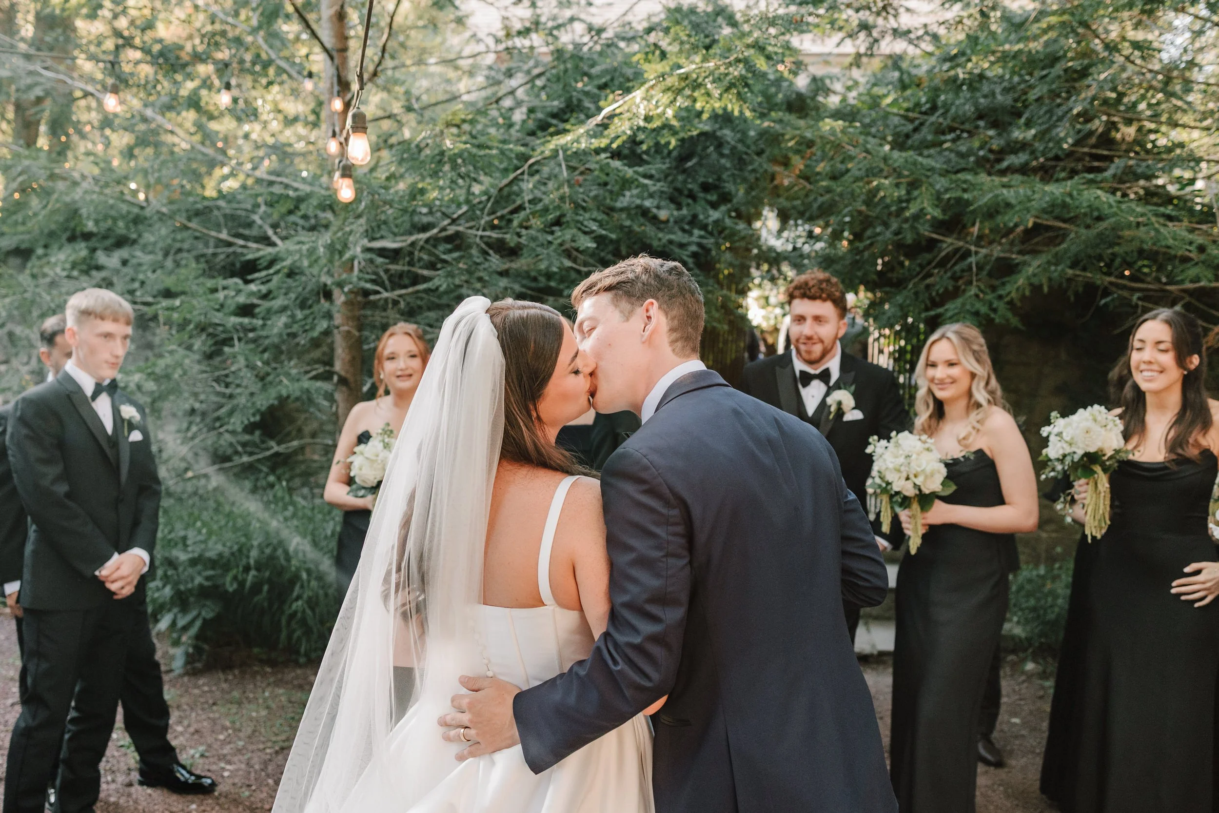 A bride and groom kiss during their outdoor wedding, with bridesmaids and groomsmen watching and smiling.