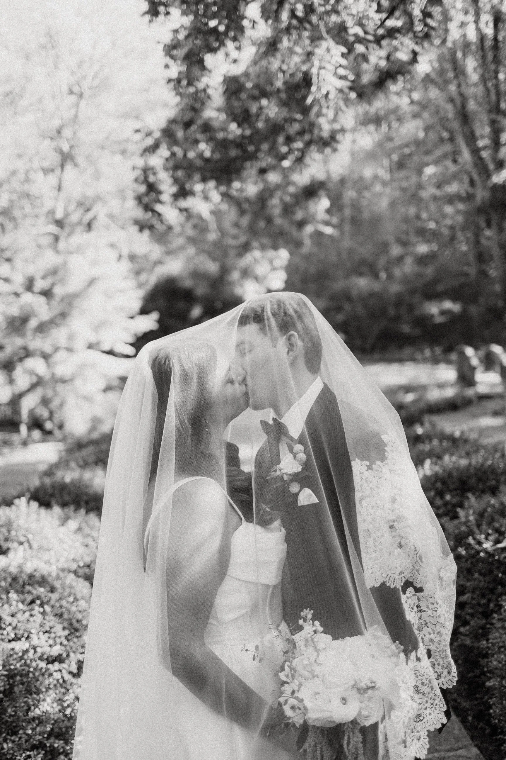 A black and white photo of a bride and groom sharing a kiss under a veil in an outdoor garden.