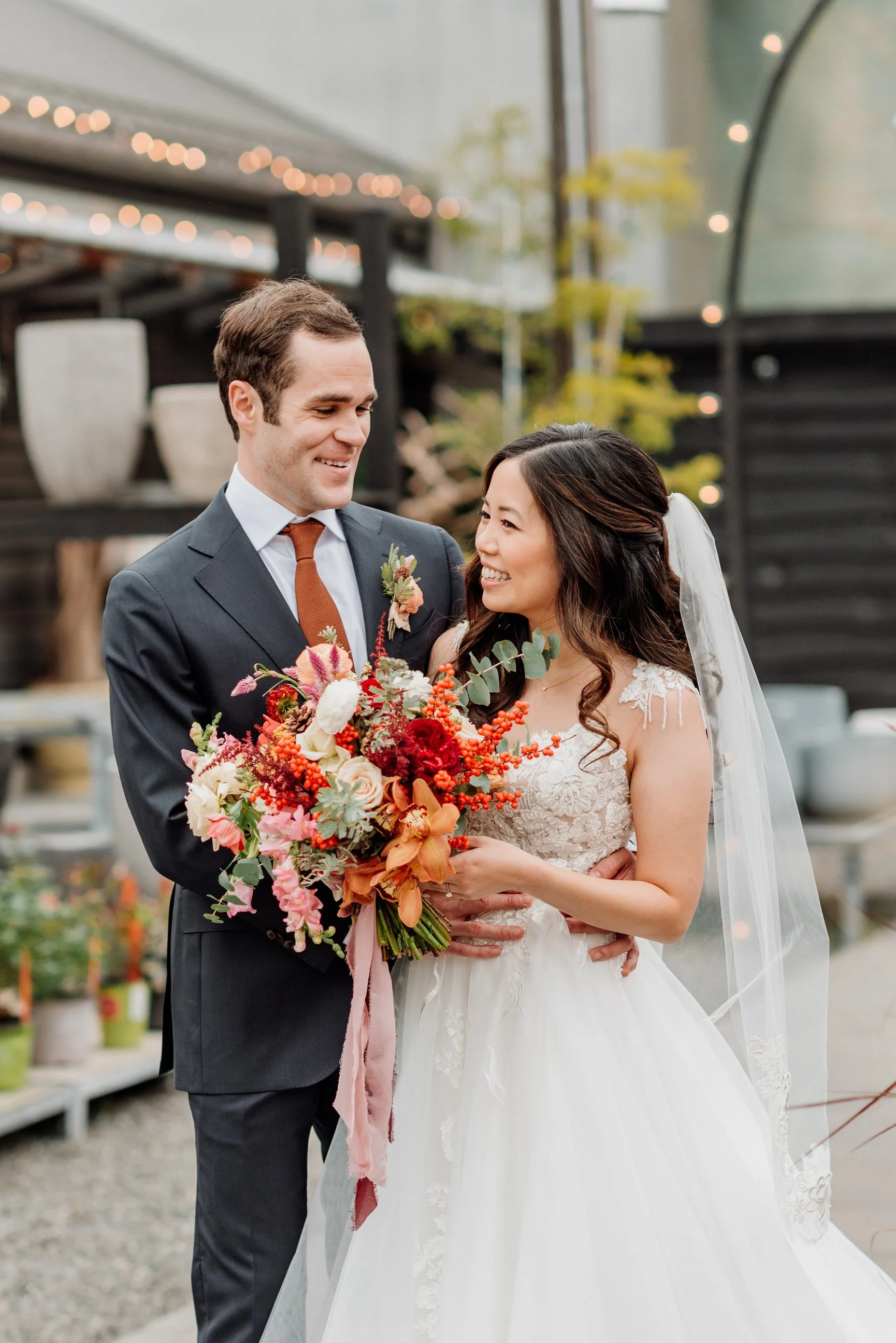 A happy couple on their wedding day, with the groom in a dark suit and tie, and the bride in a white wedding dress with lace details, holding a colorful bouquet of flowers, standing outdoors with blurred background and string lights.