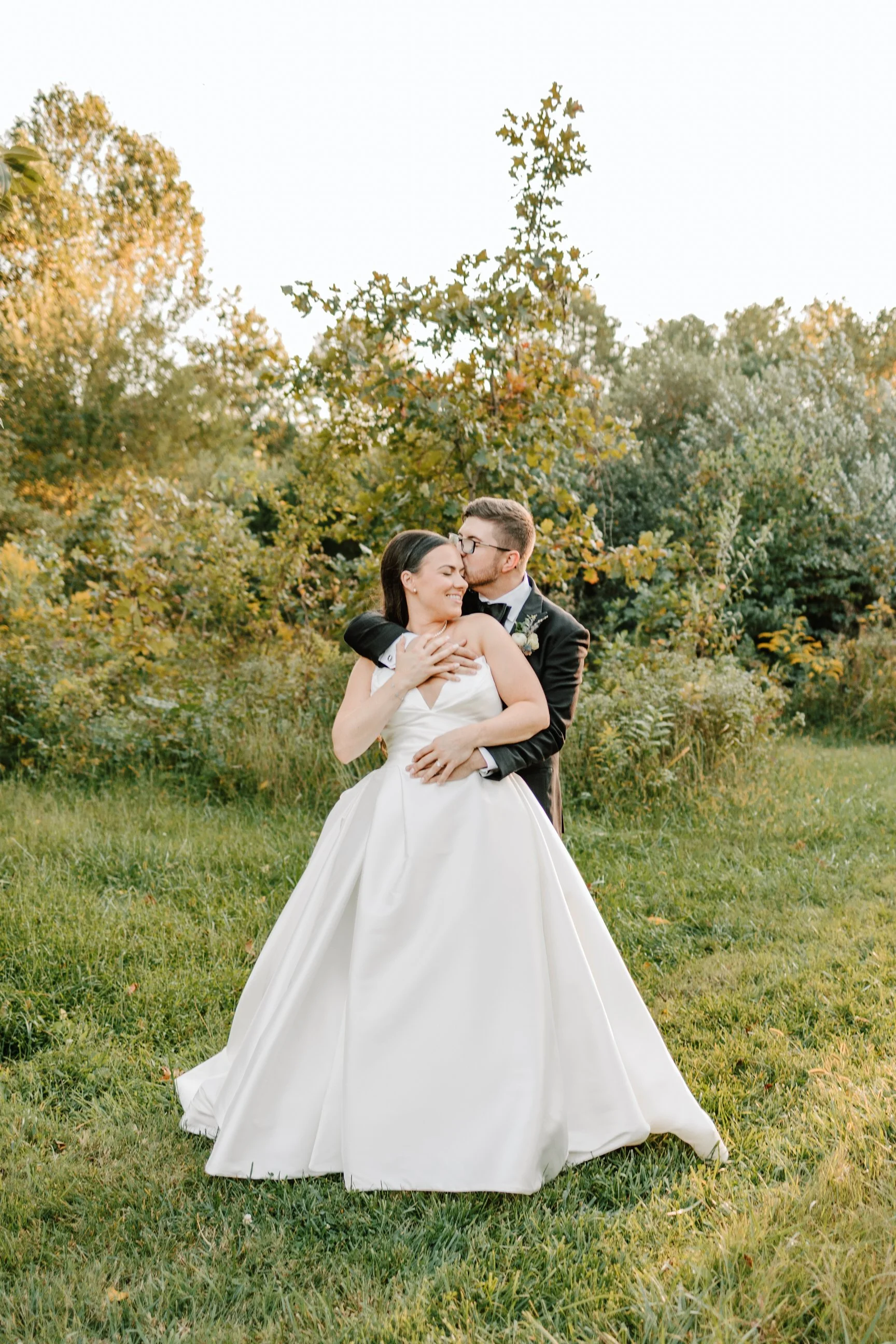 A newlywed couple in wedding attire embracing outdoors during daytime, with greenery and trees in the background.