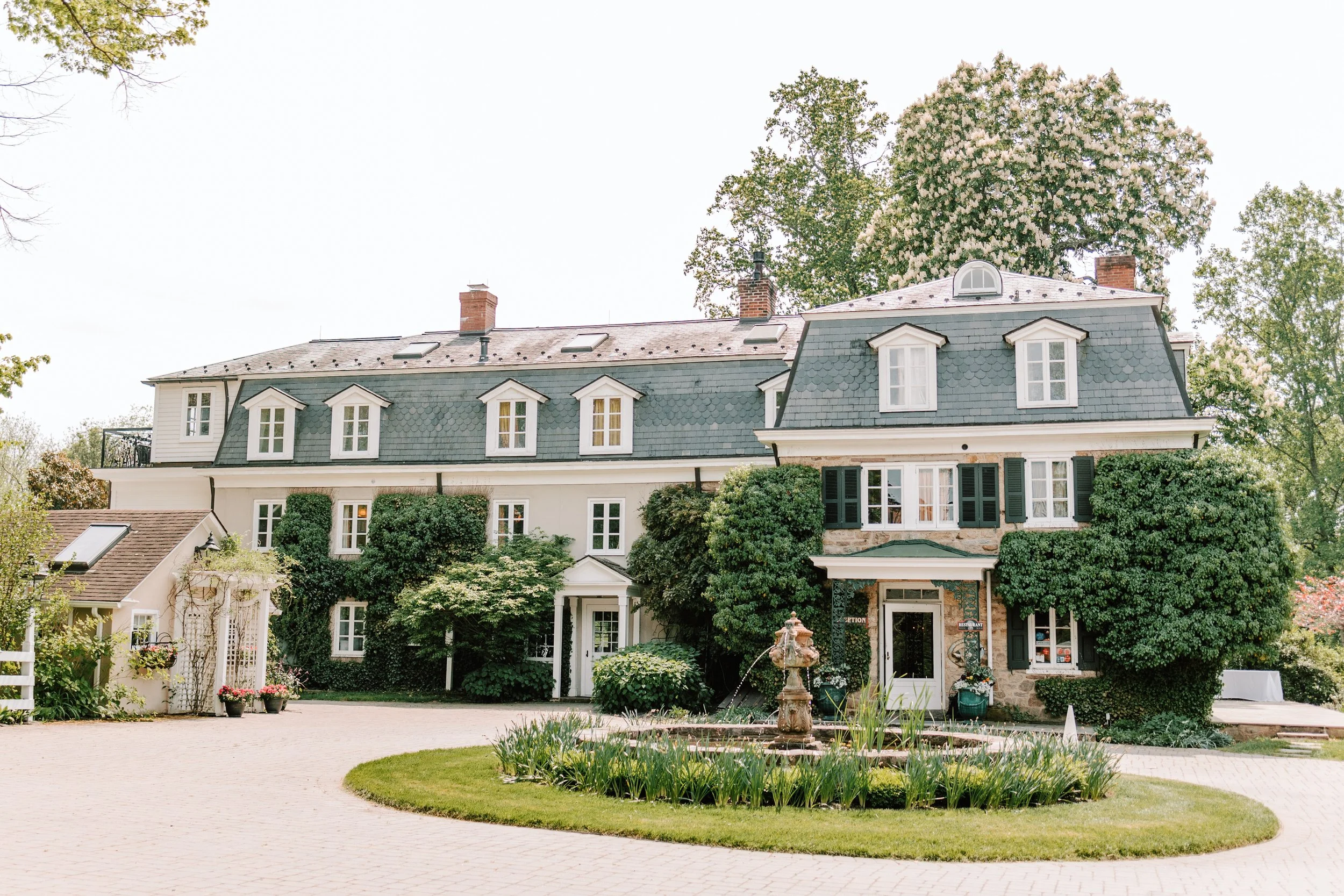 Exterior of a large, multi-story house with a mansard roof, surrounded by greenery, a fountain in the front yard, and a paved driveway.