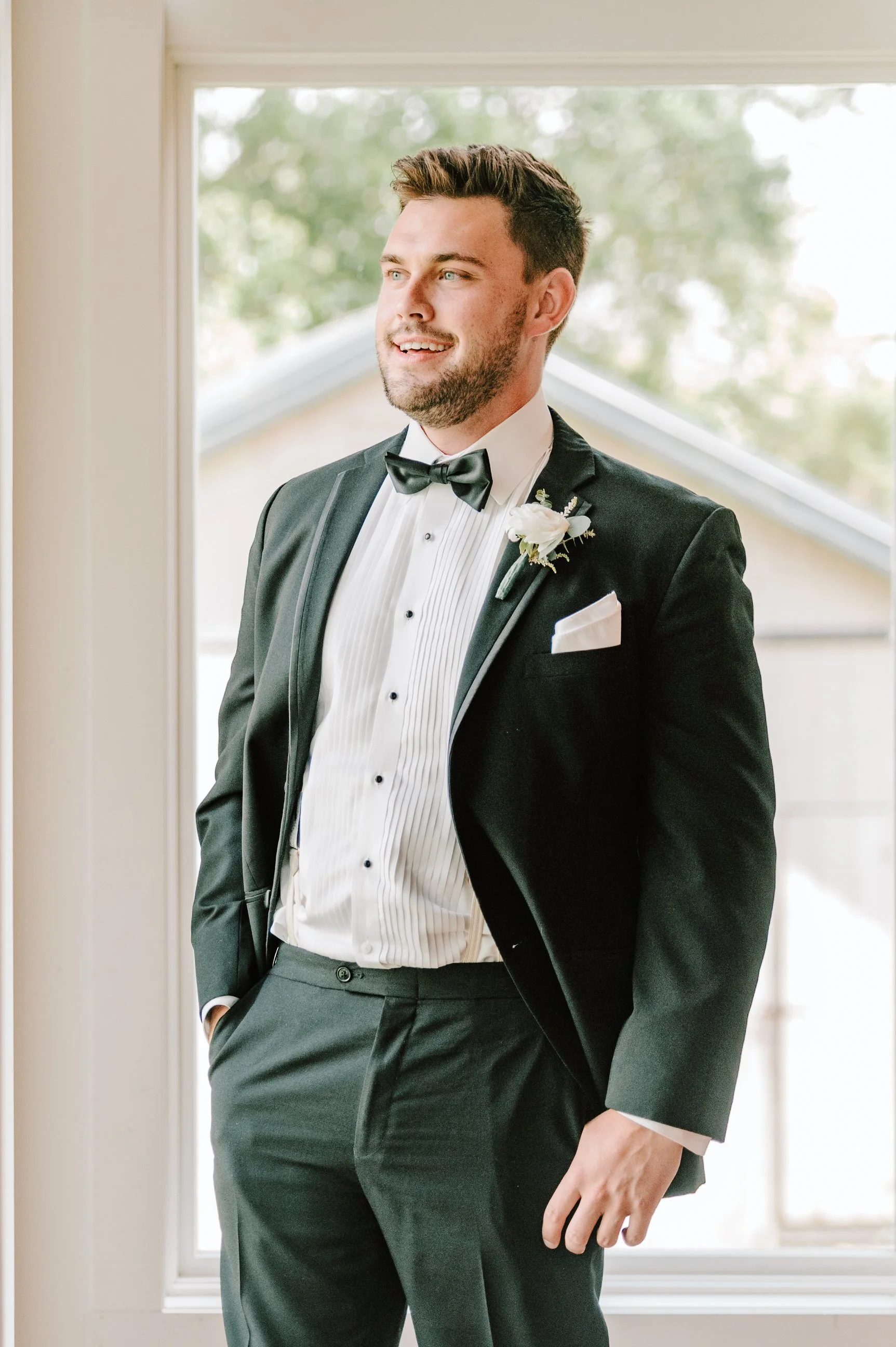 A groom standing inside, wearing a black tuxedo with a white shirt, black bow tie, and a white boutonniere, smiling and gazing outside the window.