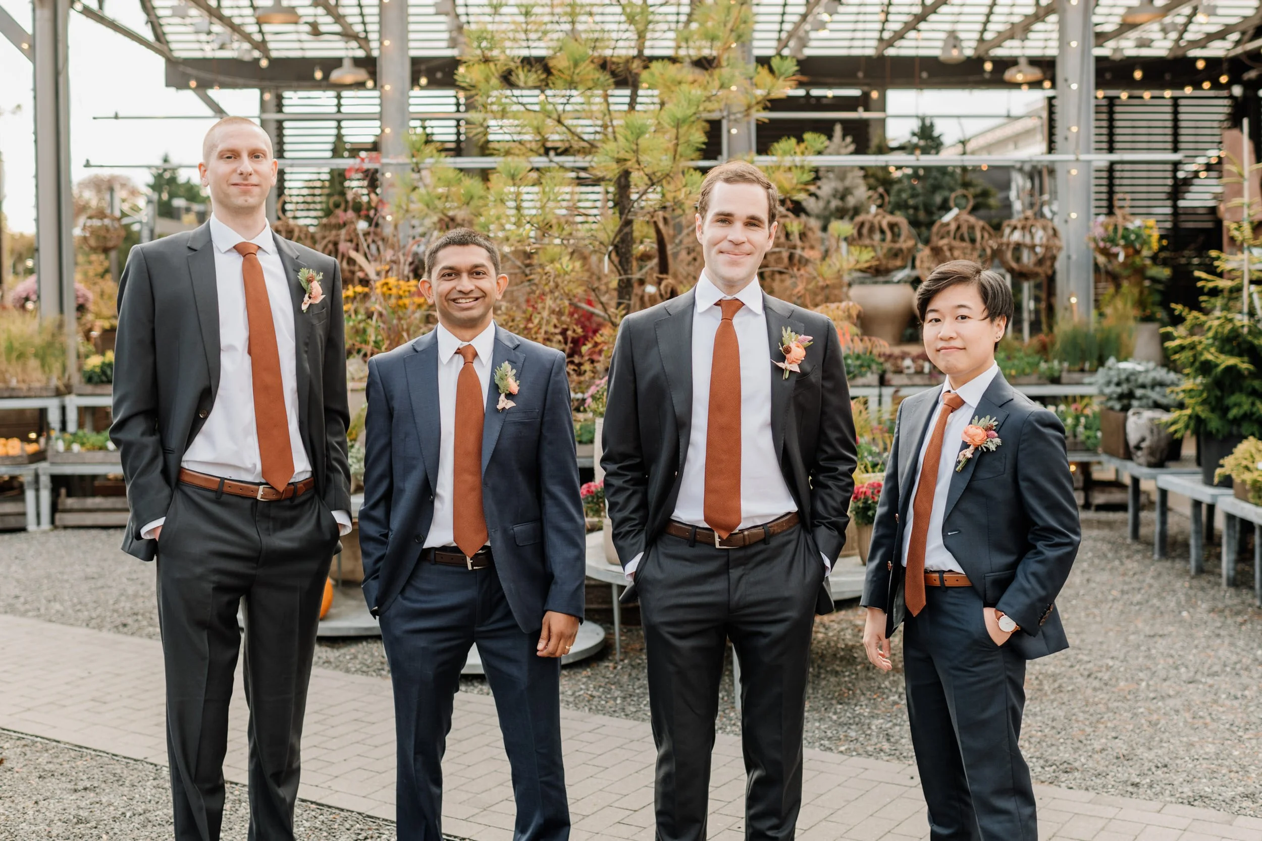 Five men in suits with orange ties and floral boutonnieres standing outdoors in a garden or greenhouse setting.