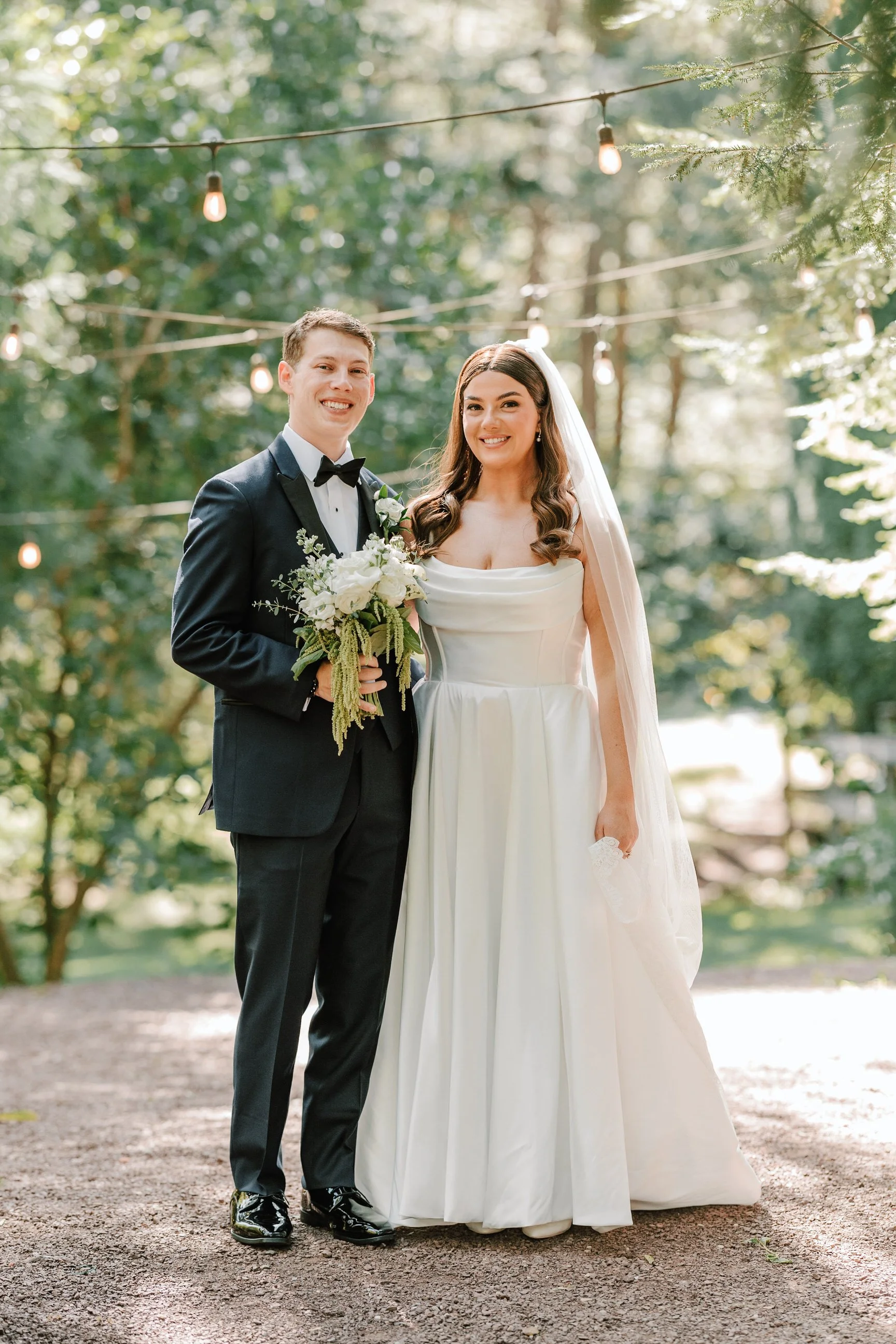 A bride and groom stand outdoors in a wooded area, smiling at the camera. The groom is wearing a black tuxedo with a bow tie, and the bride is dressed in a white wedding gown with a long veil. The bride is holding a bouquet of white flowers and green