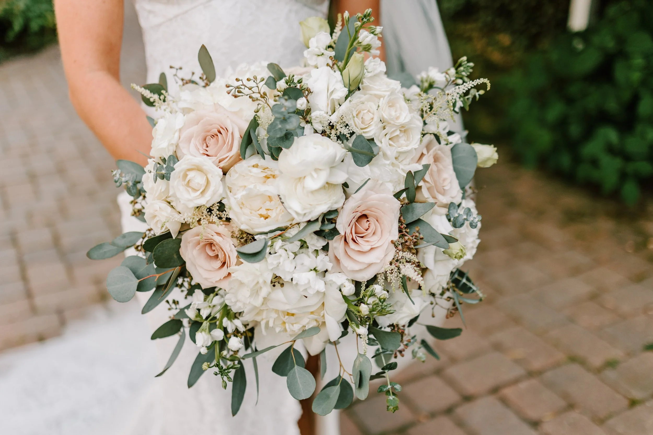 Close-up of a bridal bouquet featuring white and blush pink roses, peonies, and greenery, held by a bride in a wedding dress.