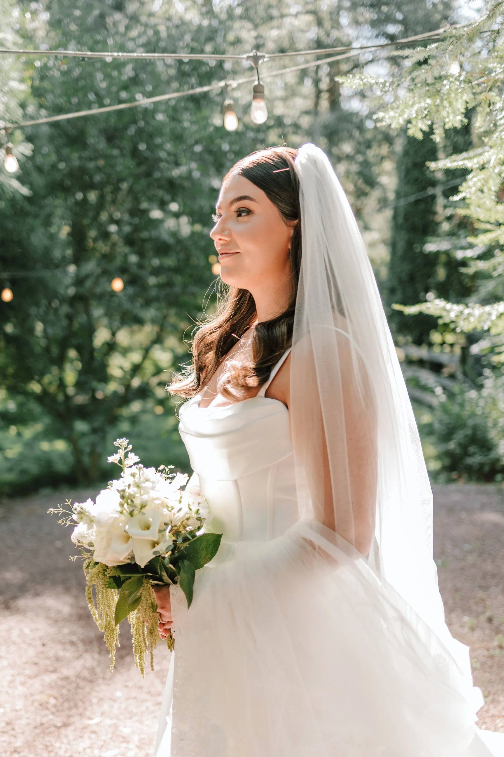 A bride in a white wedding dress with a veil holding a bouquet of white flowers outdoors with trees and string lights overhead.