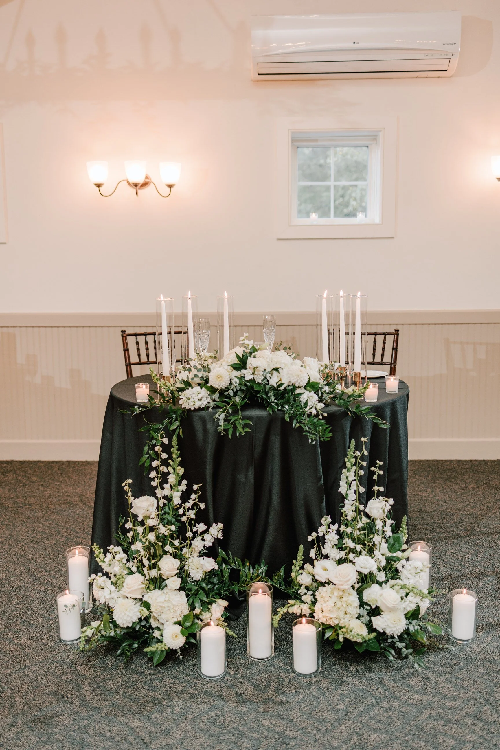 Elegant rectangular table with black tablecloth, floral centerpiece with white flowers and greenery, surrounded by candles in glass holders, set in a softly lit indoor space.