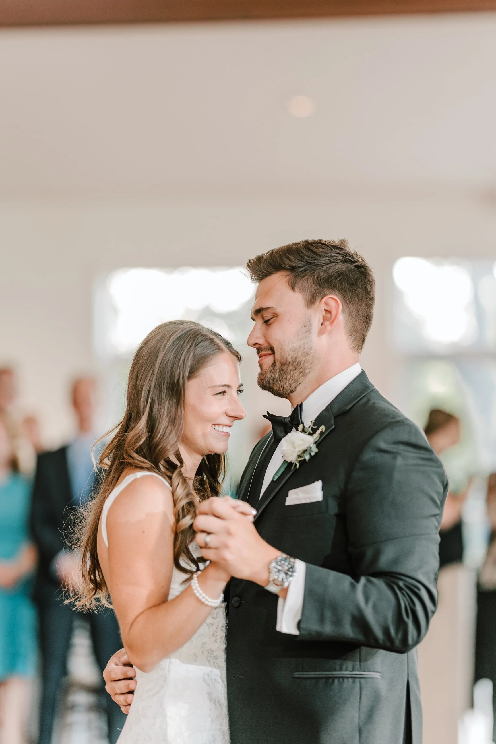 A bride and groom during their wedding dance, smiling and looking at each other, with guests blurred in the background.
