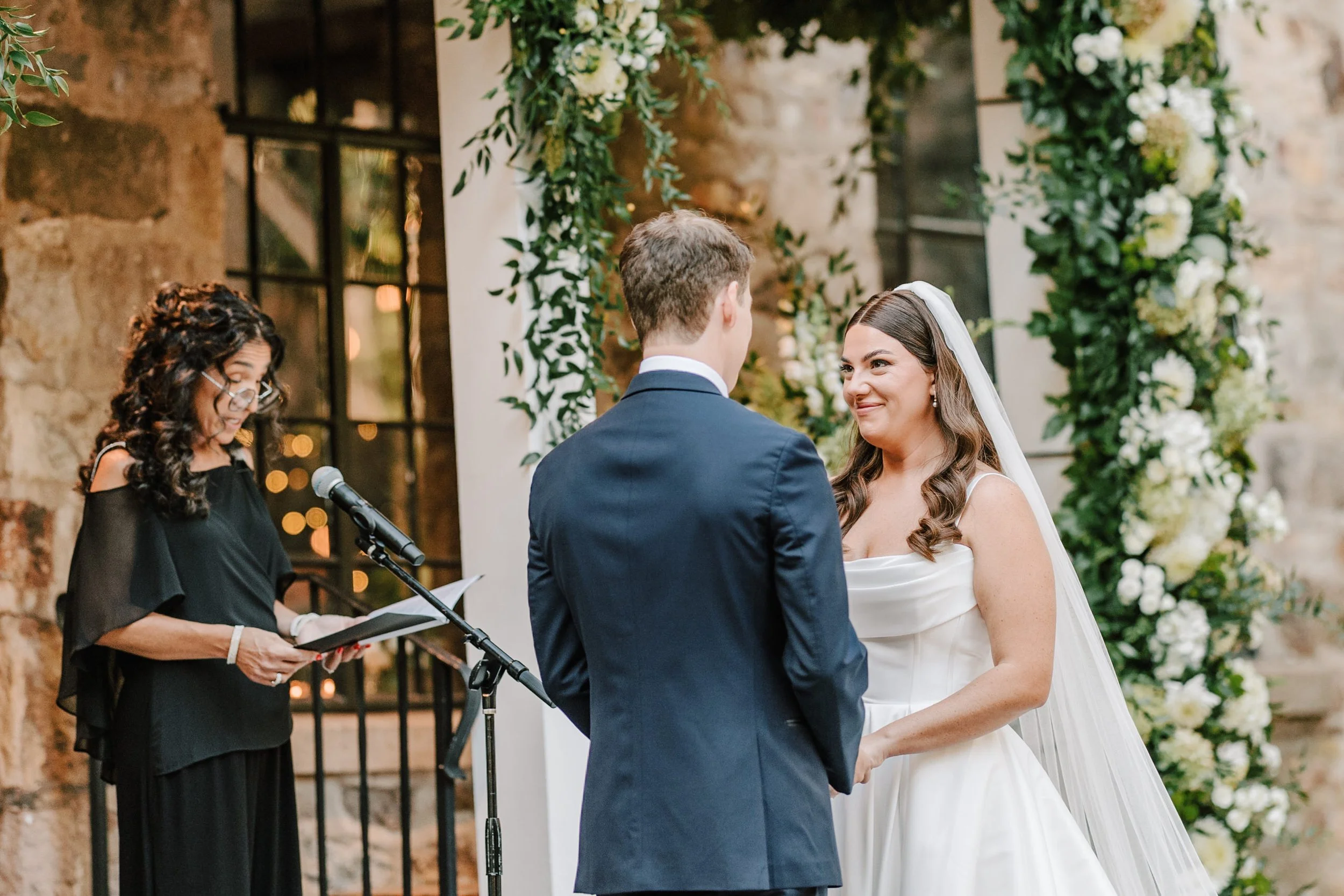 A bride and groom exchange vows at their wedding ceremony, standing in front of an officiant reading from a book, with a floral arch in the background.