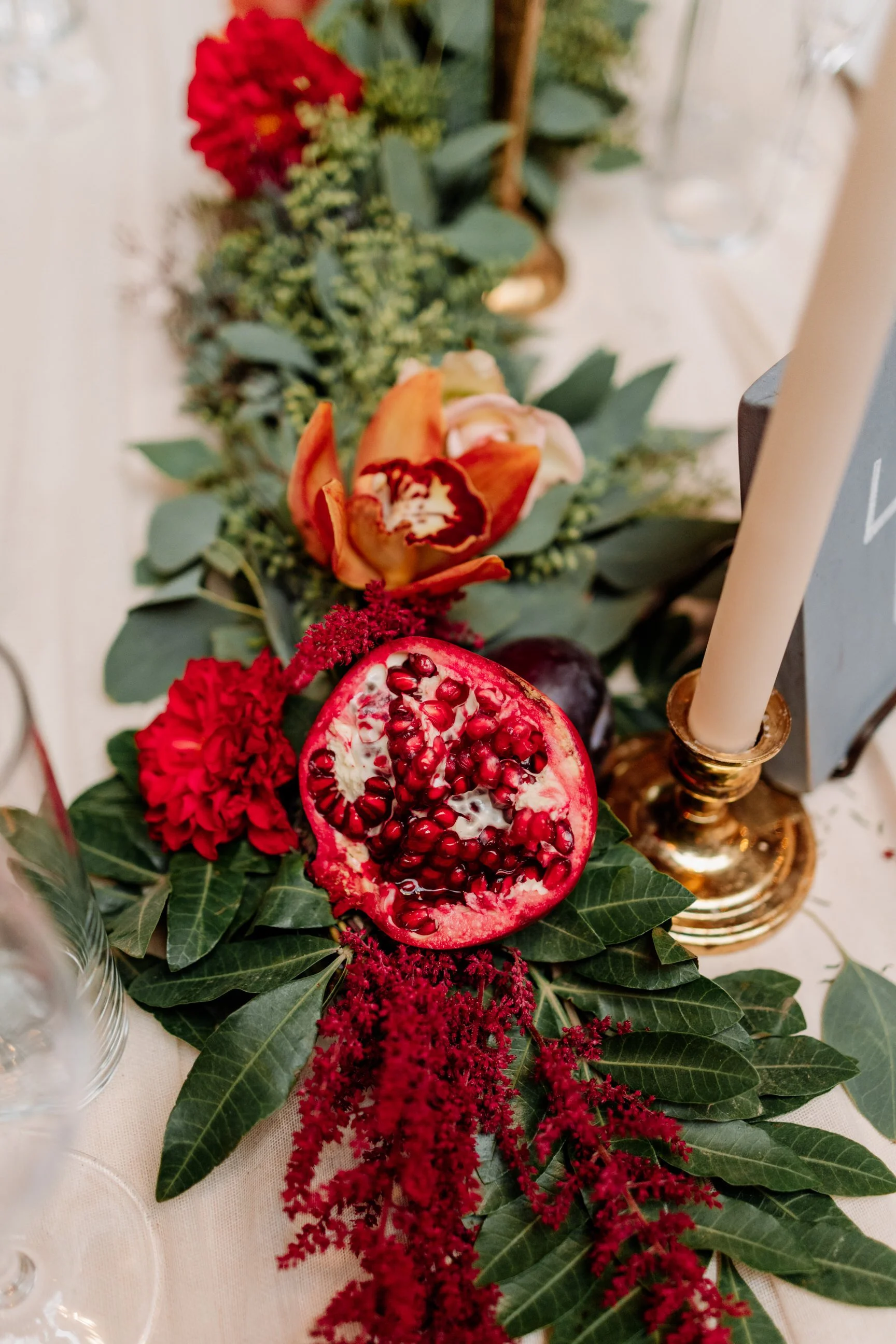 A centerpiece featuring a pomegranate, red carnations, orange tulips, greenery, and a gold candlestick with a white candle.