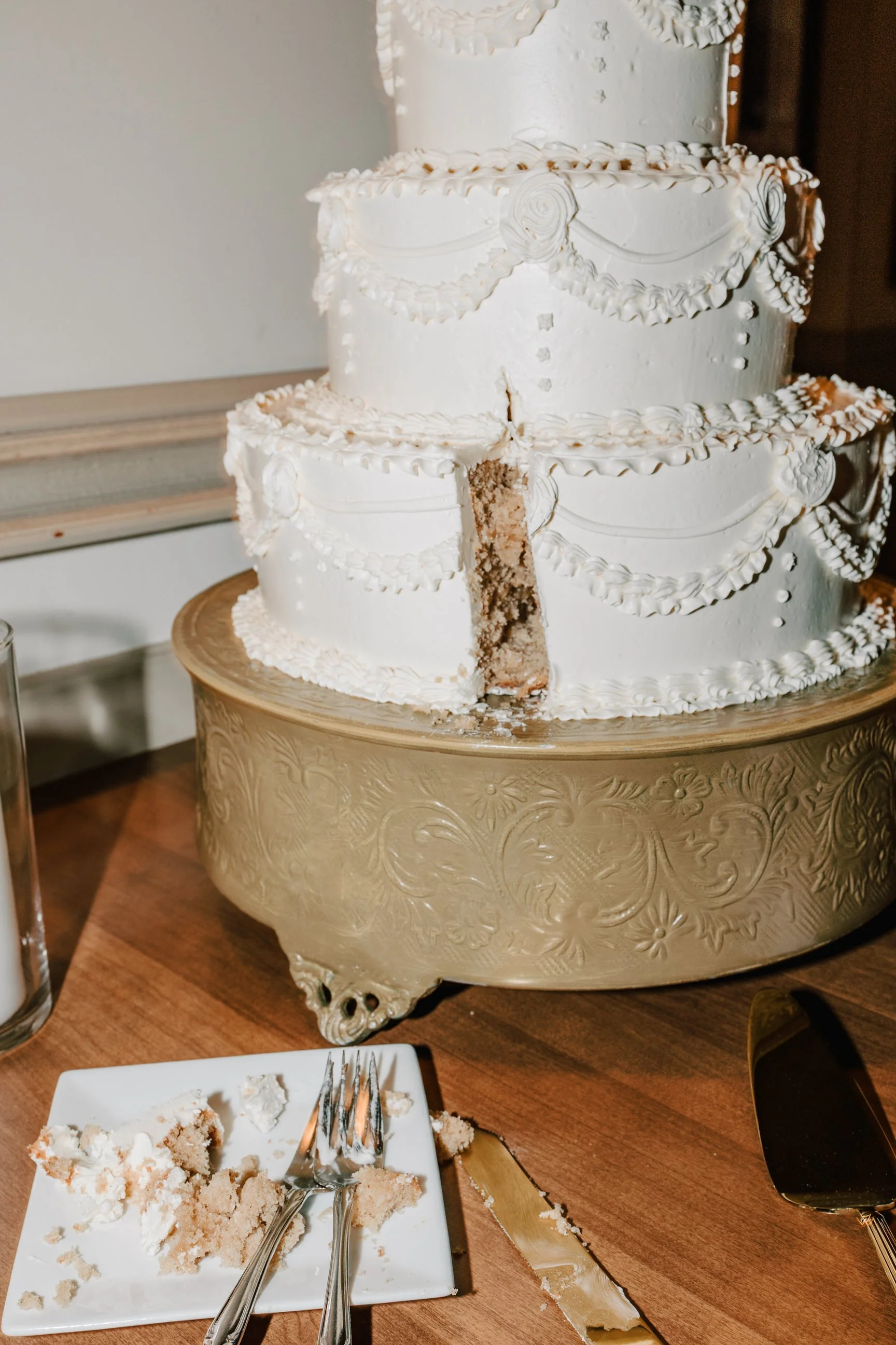 A three-tiered white wedding cake with decorative icing borders and drapes, partially cut with a slice and crumbs on a plate, sitting on a wooden table with a gold cake stand.
