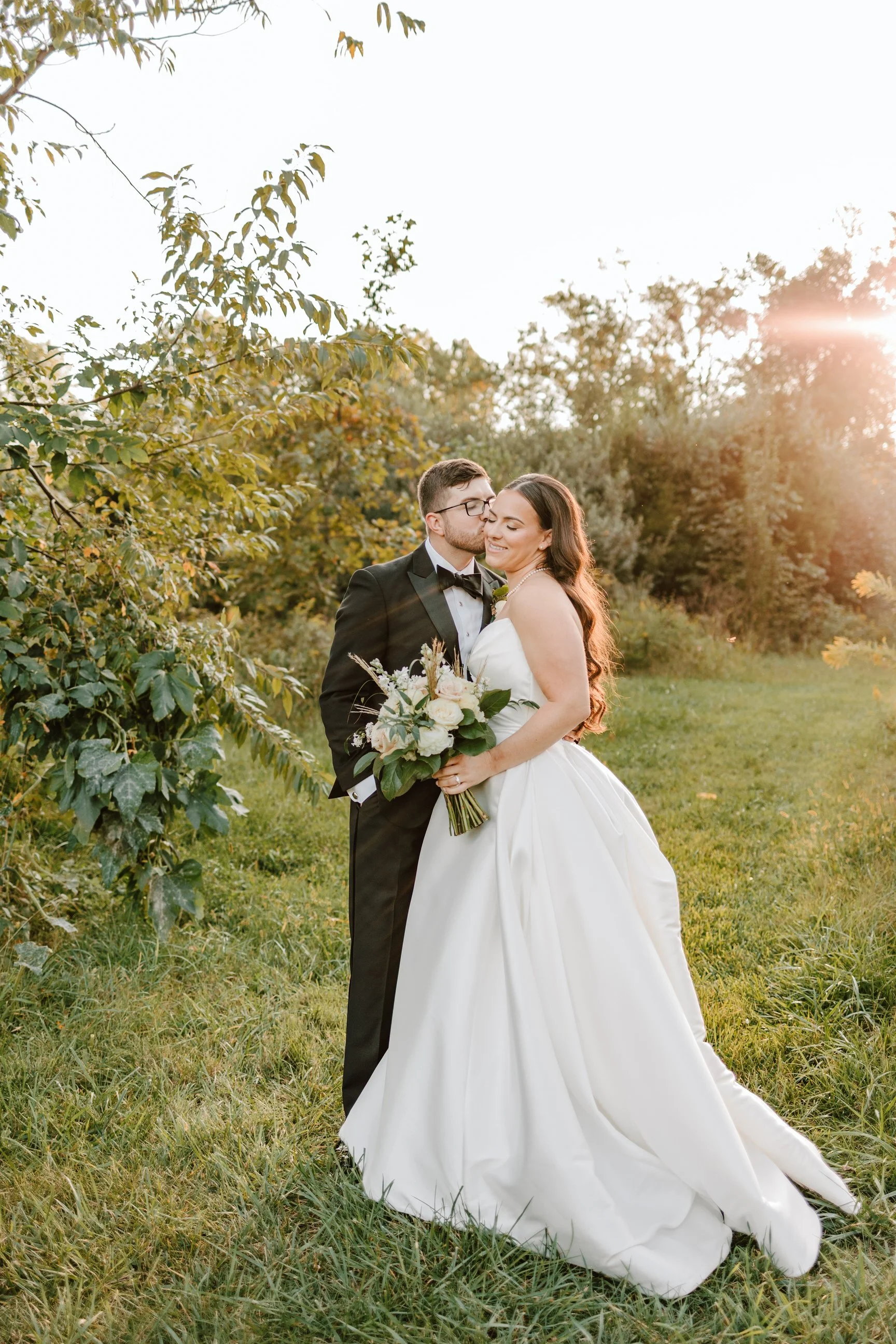A newlywed couple standing close together outdoors during sunset, with the groom in a black tuxedo kissing the bride's forehead as she smiles, holding a bouquet of white and pink flowers.