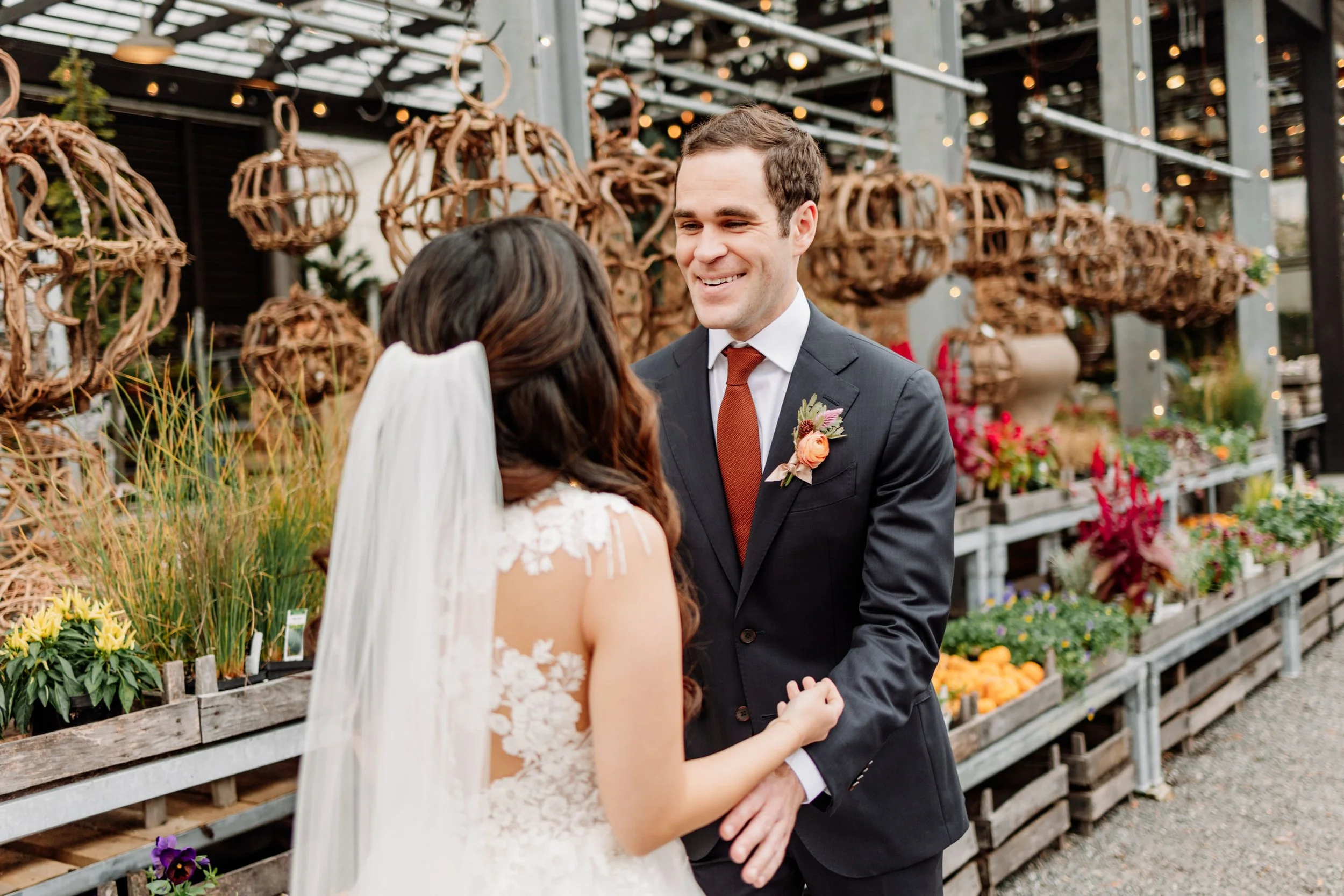 A bride and groom holding hands while exchanging vows outdoors at a wedding, with a backdrop of plants and decorative wicker balls.