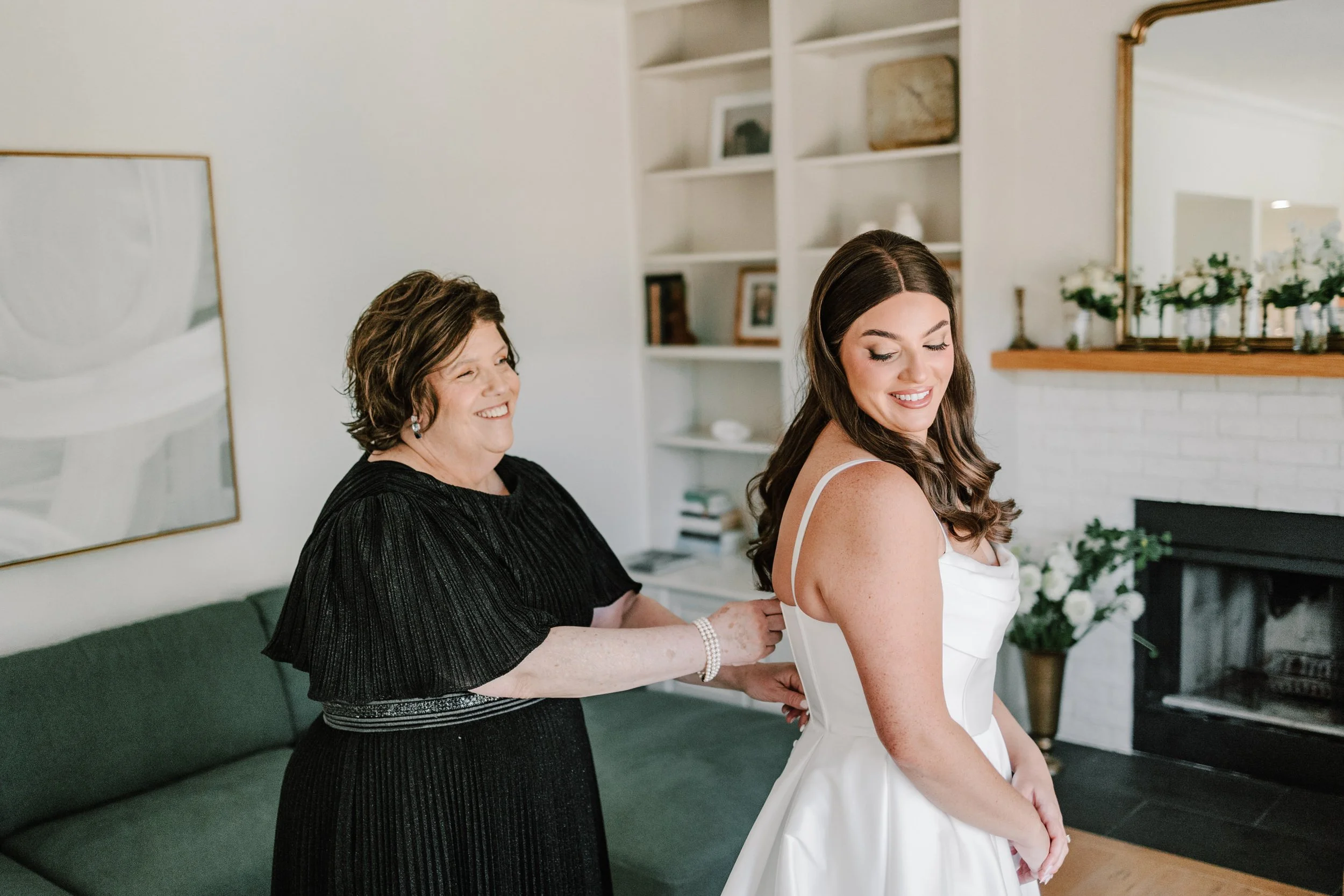 A young woman in a white dress smiling as she adjusts her dress, with an older woman in a black dress helping her from behind in a bright living room.