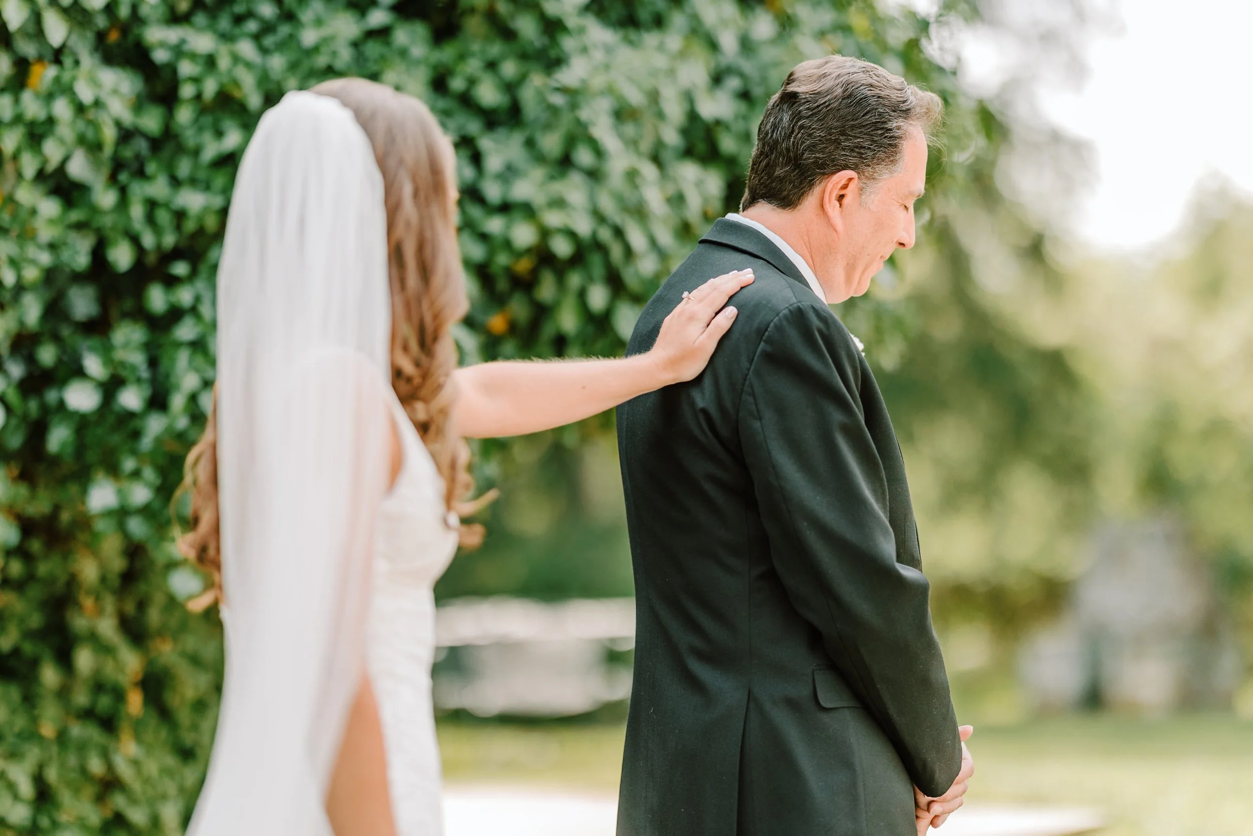 A bride gently touching her father on the shoulder during a wedding ceremony outdoors.
