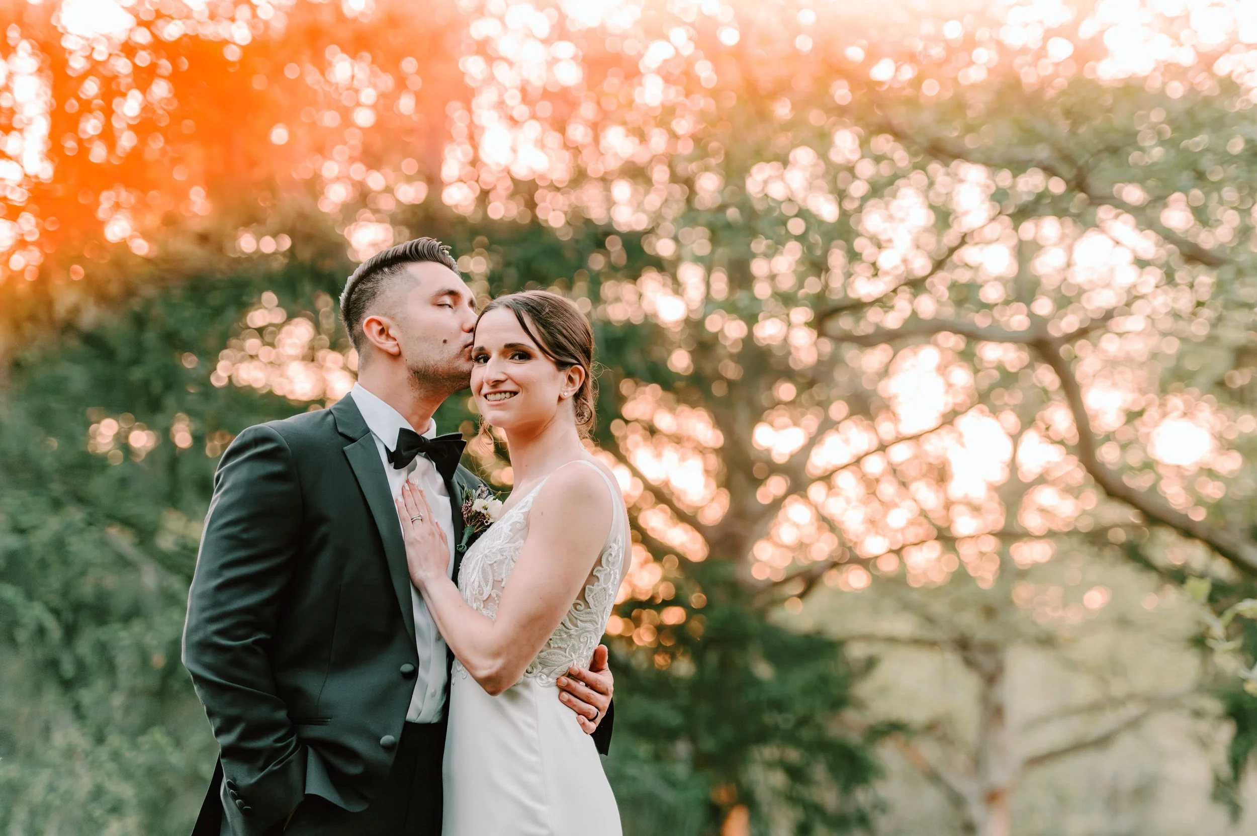 A newlywed couple in wedding attire standing outdoors with trees and sunset in the background. The groom is kissing the bride on the temple while she smiles at the camera.