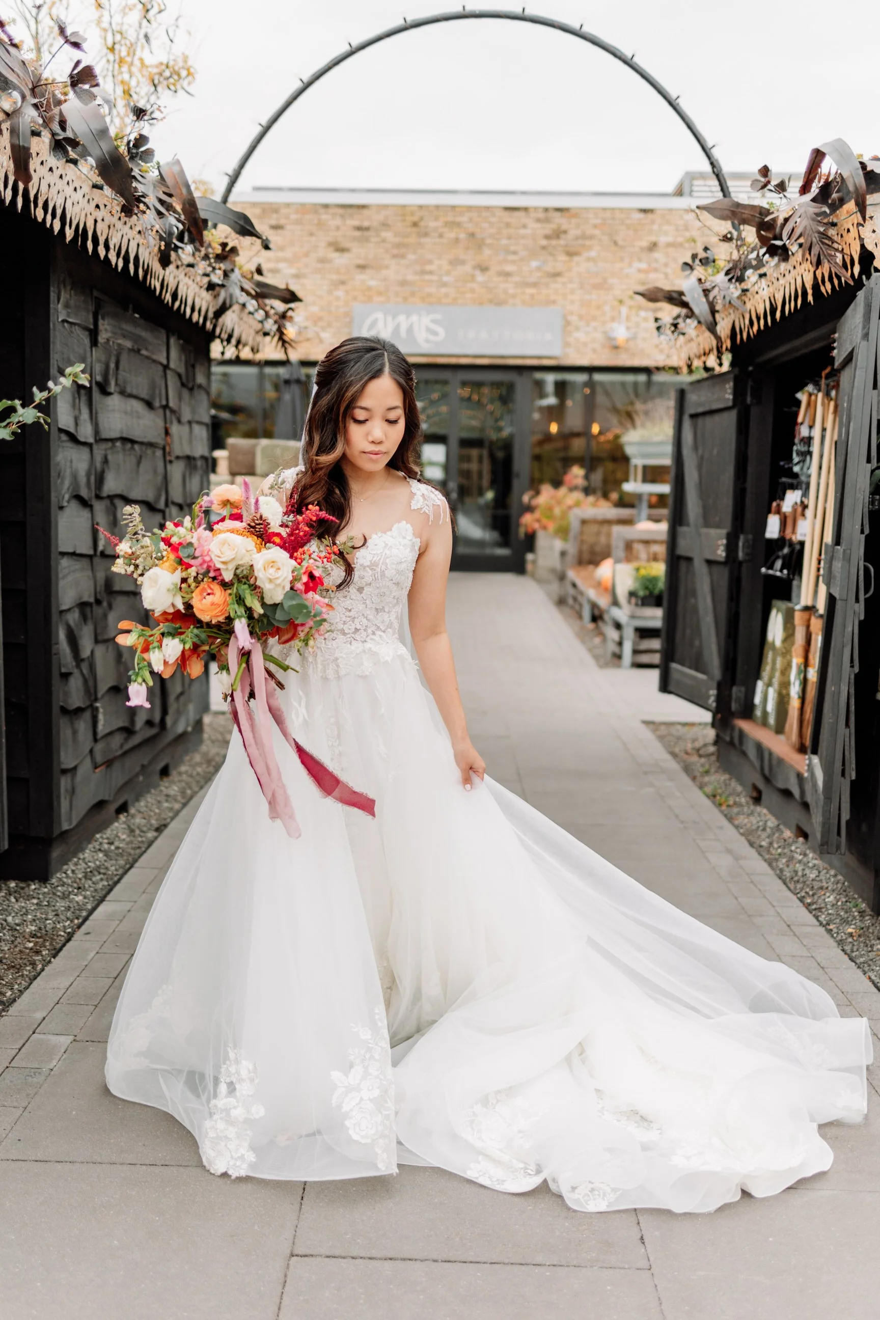 A bride in a white wedding dress holding a bouquet of colorful flowers, standing outdoors on a paved walkway with black wooden fencing and an archway