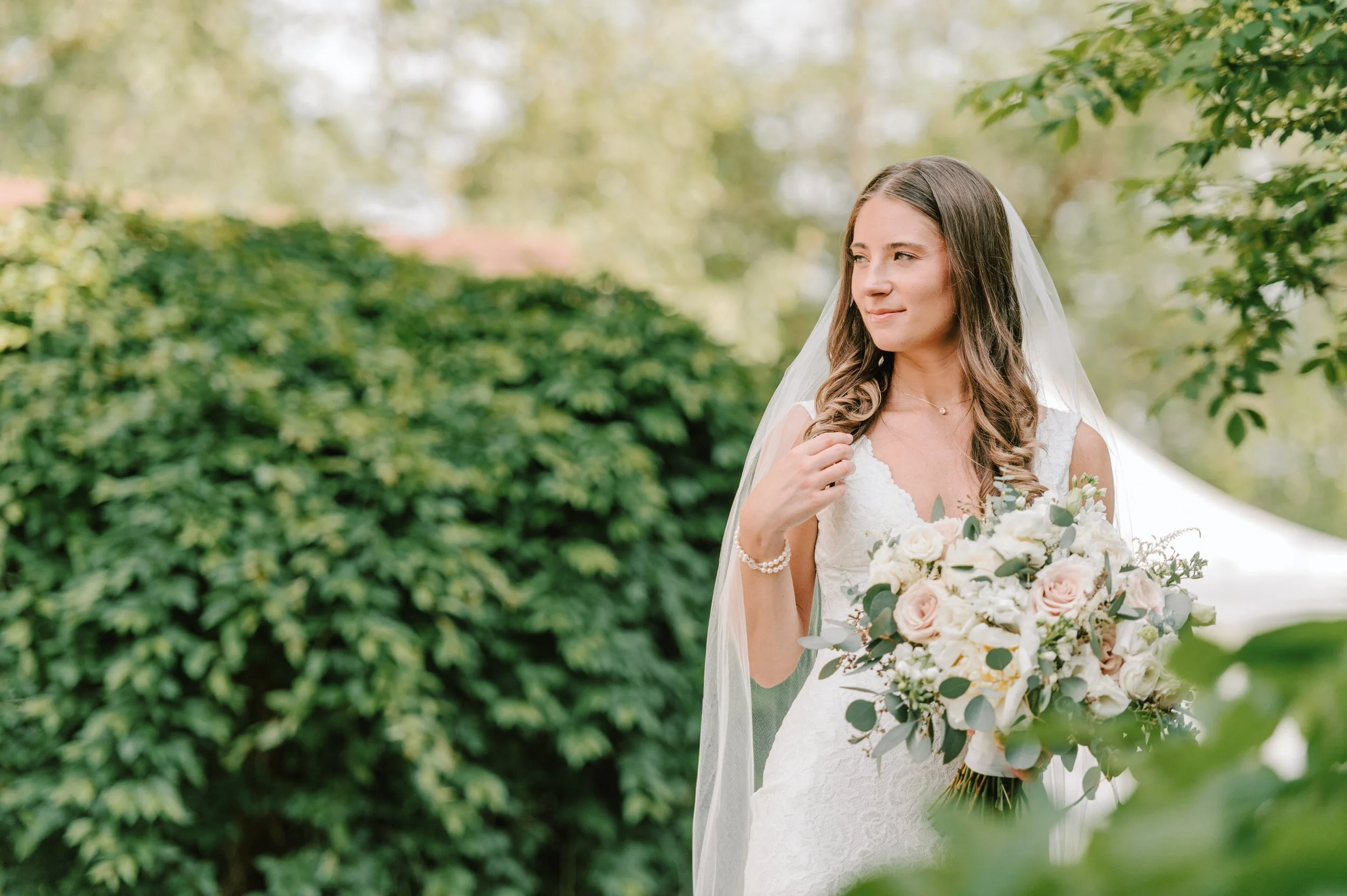 A bride holding a bouquet of white and blush pink flowers, standing outdoors with greenery in the background, wearing a lace wedding dress and a veil.