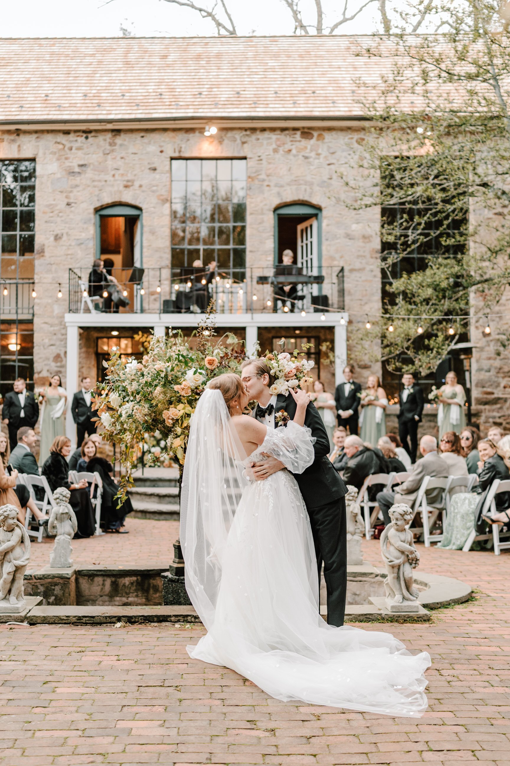 A bride and groom kiss during their wedding ceremony in an outdoor courtyard with a stone building in the background. The bride wears a white wedding dress with a long train and veil, and the groom wears a black tuxedo. Guests sit on white chairs, an