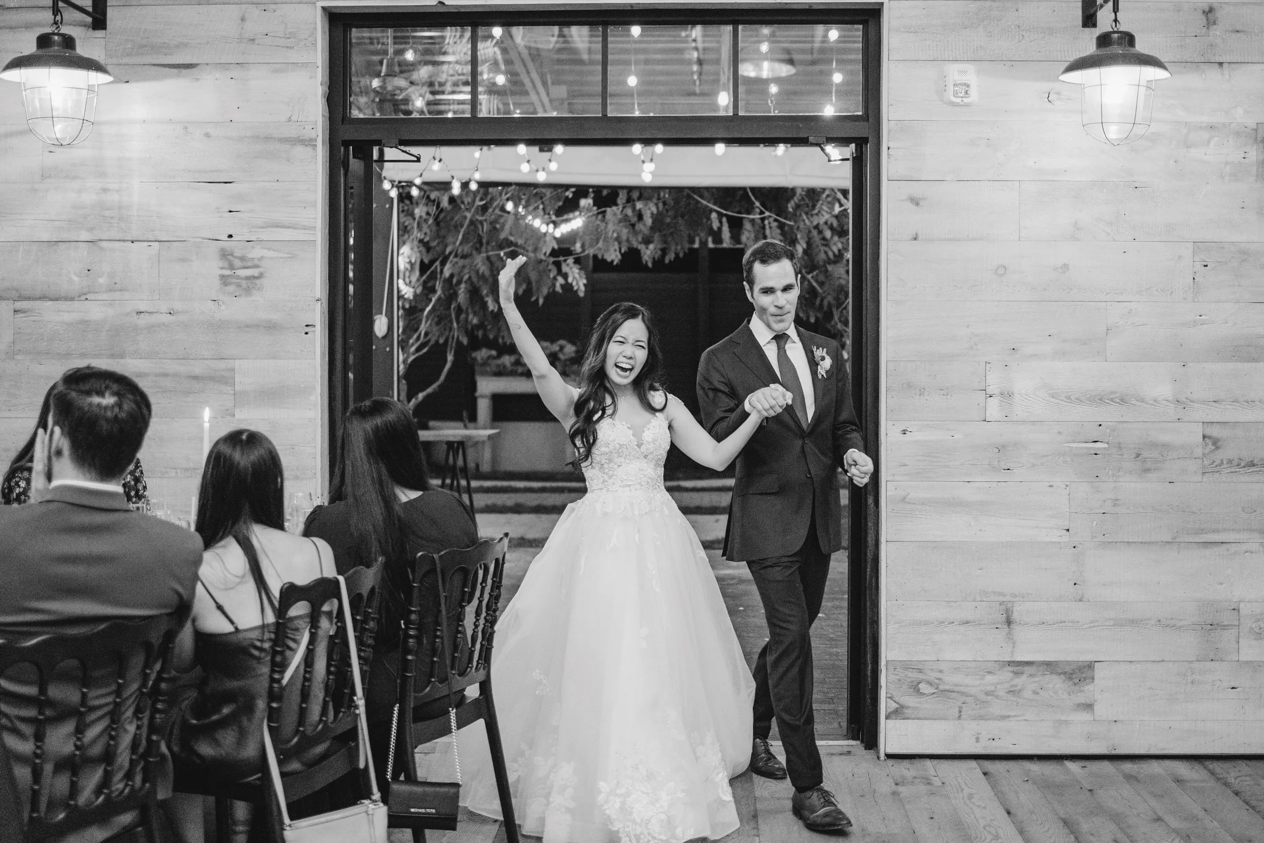 Black and white photo of a bride and groom walking into a wedding reception, holding hands, with guests seated at tables in the foreground.