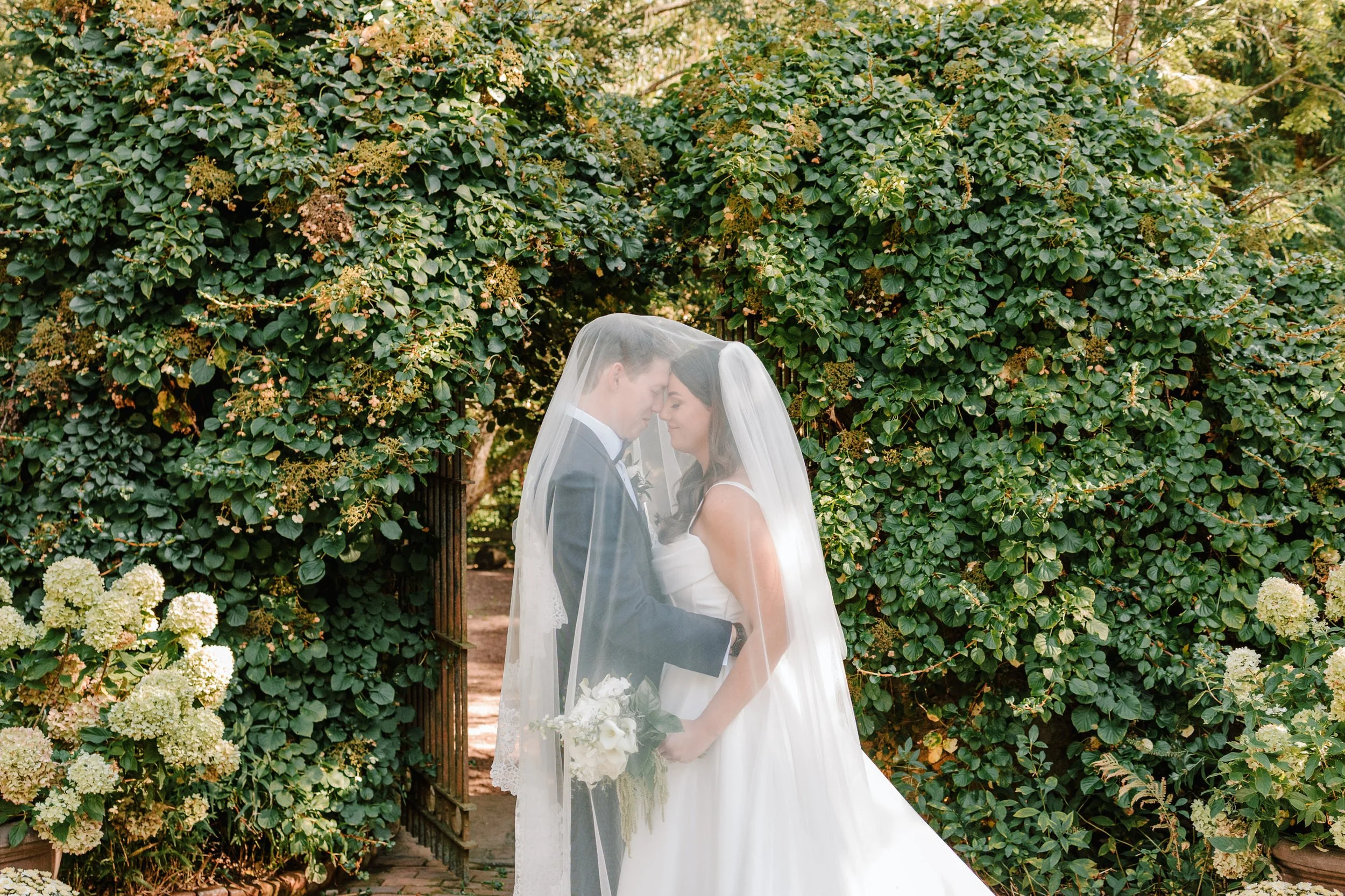 A bride and groom standing close together, with foreheads touching, under a veil, surrounded by greenery and flowers, during their wedding. Philadelphia wedding photographer