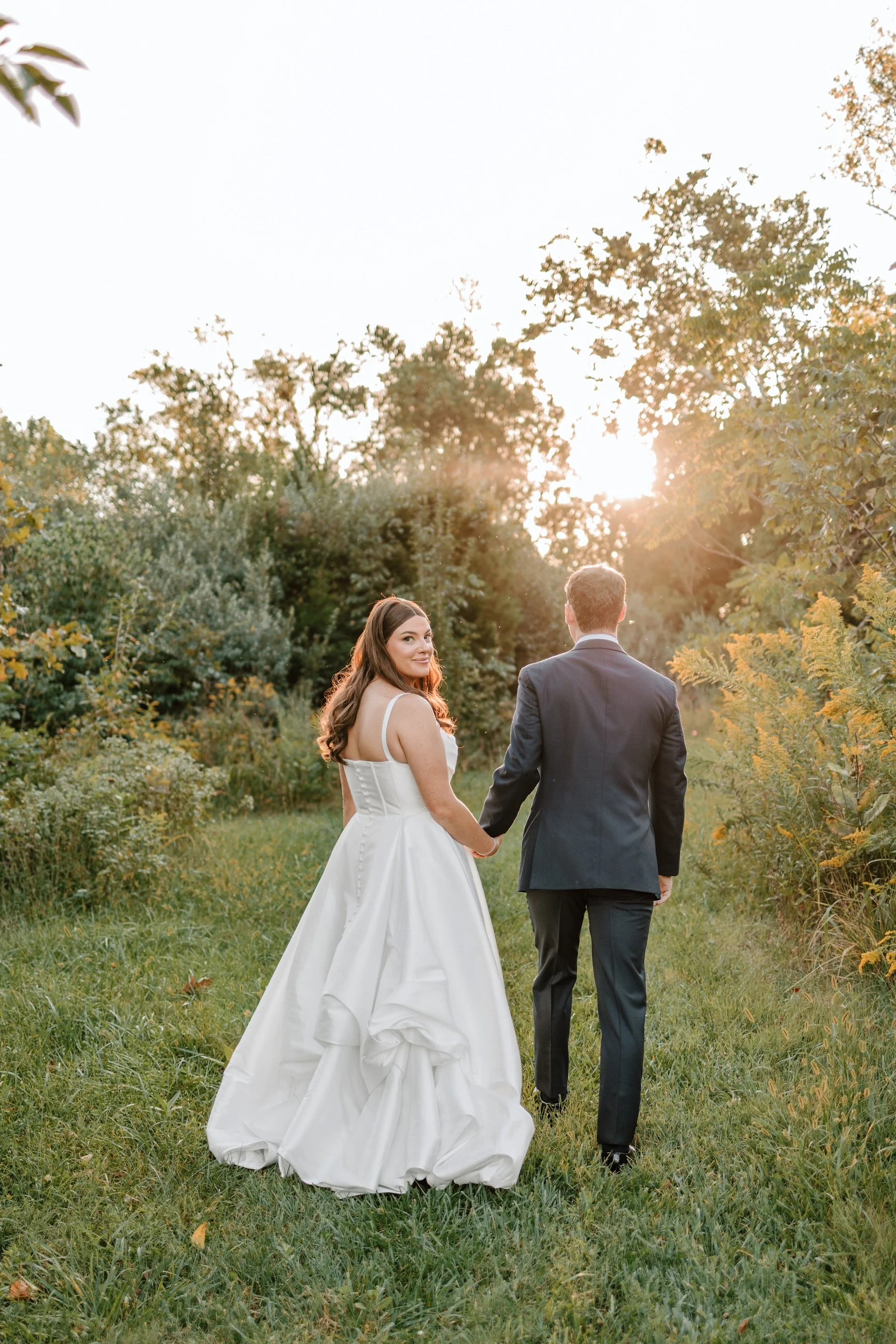 A bride and groom walking hand-in-hand on a grassy path surrounded by trees at sunset, the bride in a white wedding gown and the groom in a dark suit.