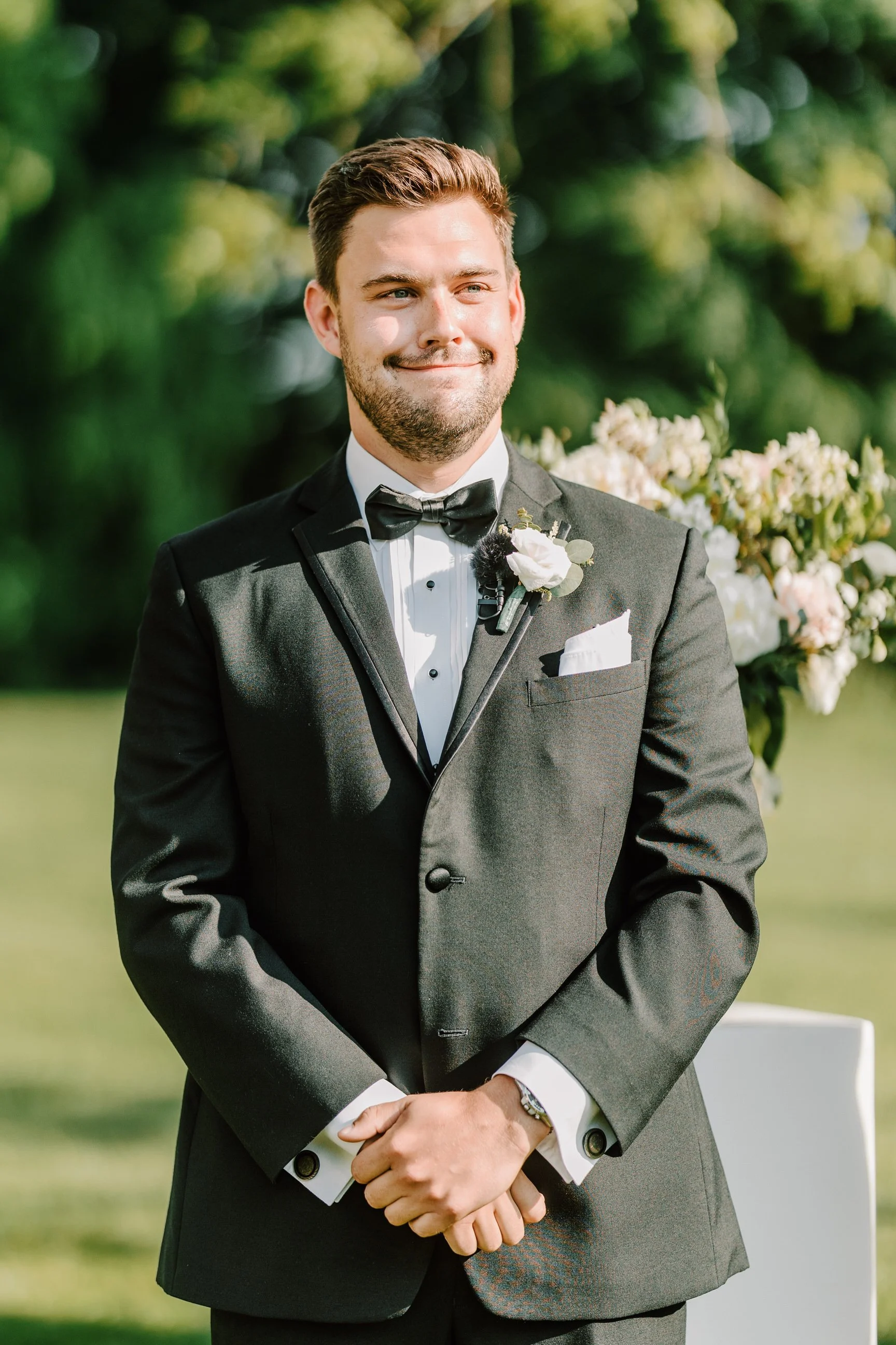 A man in a black tuxedo with a white shirt and black bow tie, standing outdoors during a wedding ceremony with a floral arrangement in the background.