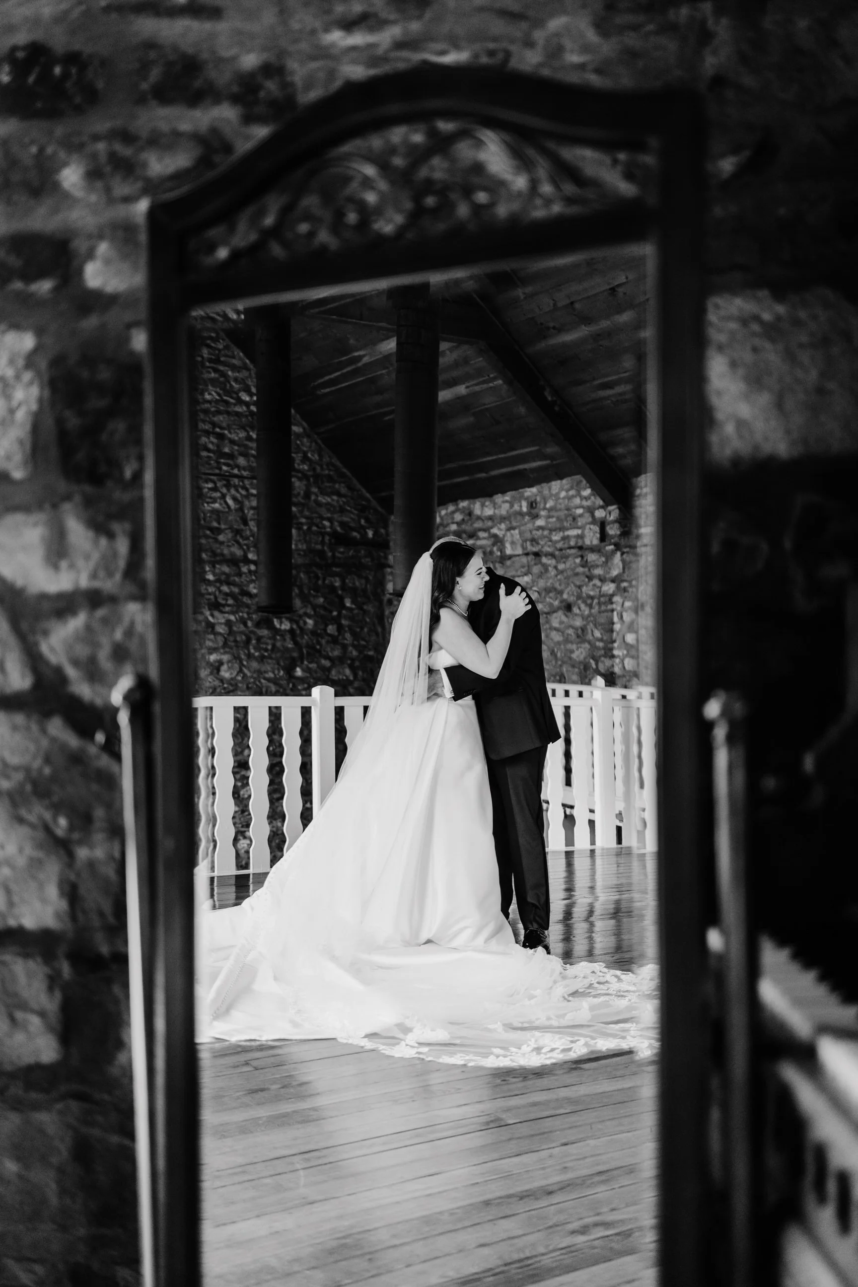 A bride and groom embrace during their wedding dance, reflected in a mirror with a stone wall and wooden ceiling in the background, in black and white.