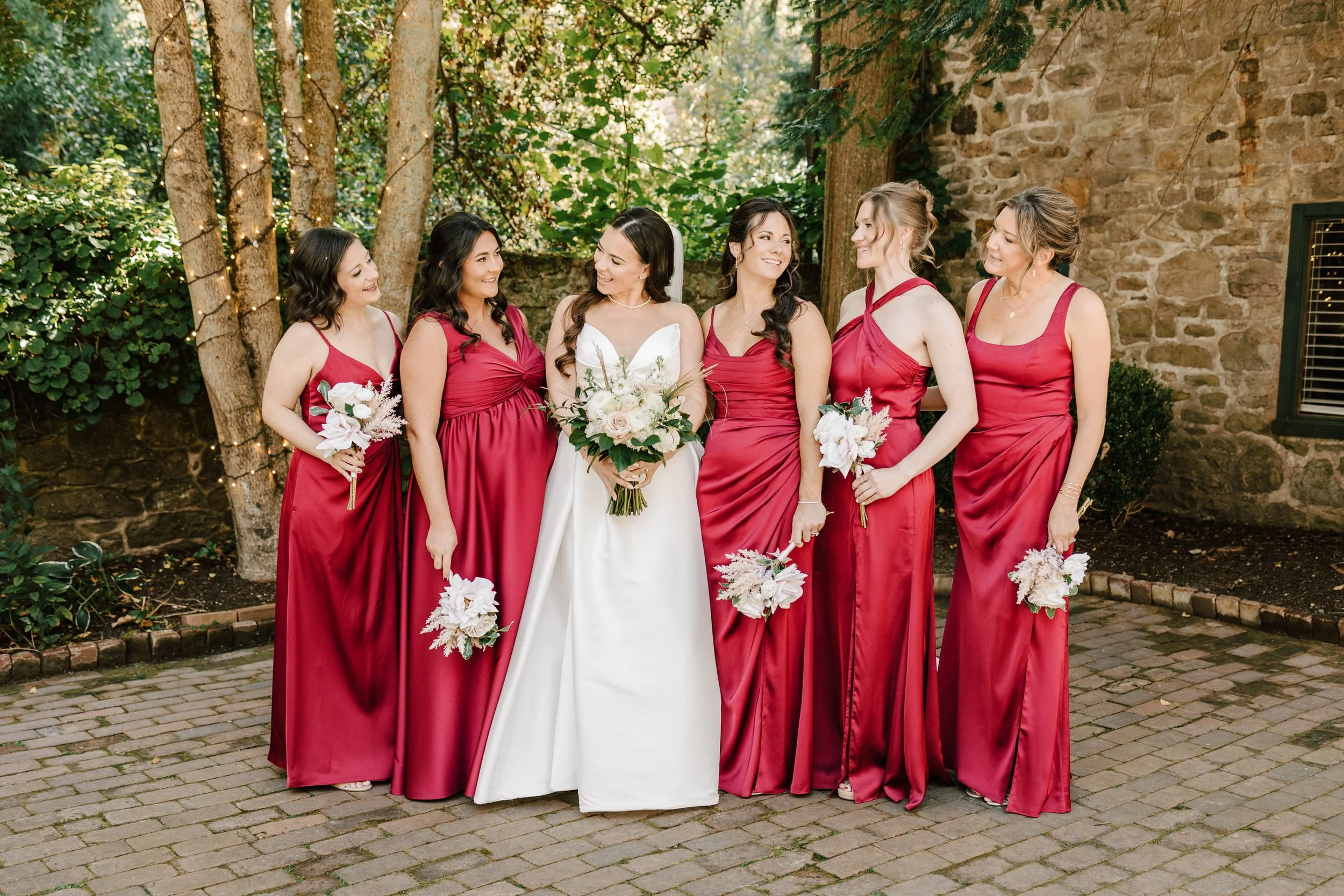 Bride in white gown with five bridesmaids in red dresses outdoors near a stone wall and trees.