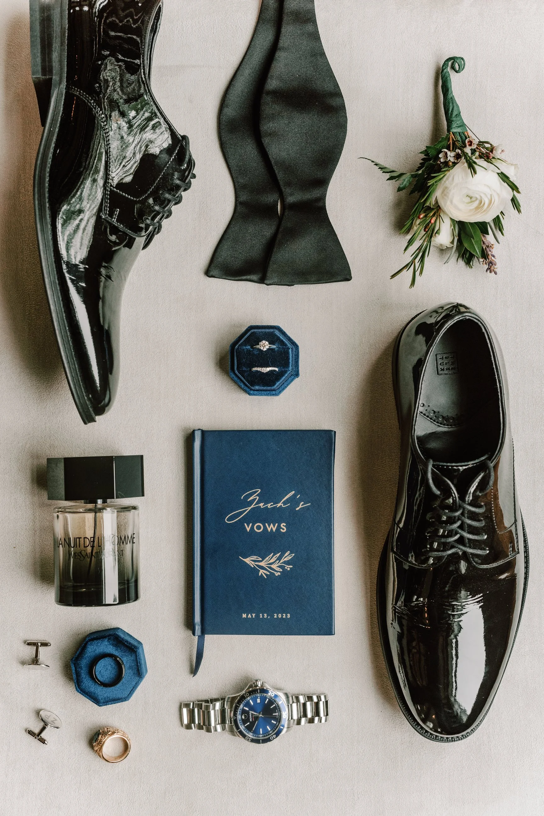 Wedding accessories including black patent leather shoes, black gloves, white flowers, jewelry, a blue vow book, perfume, a watch, and wedding rings arranged on a light-colored surface.