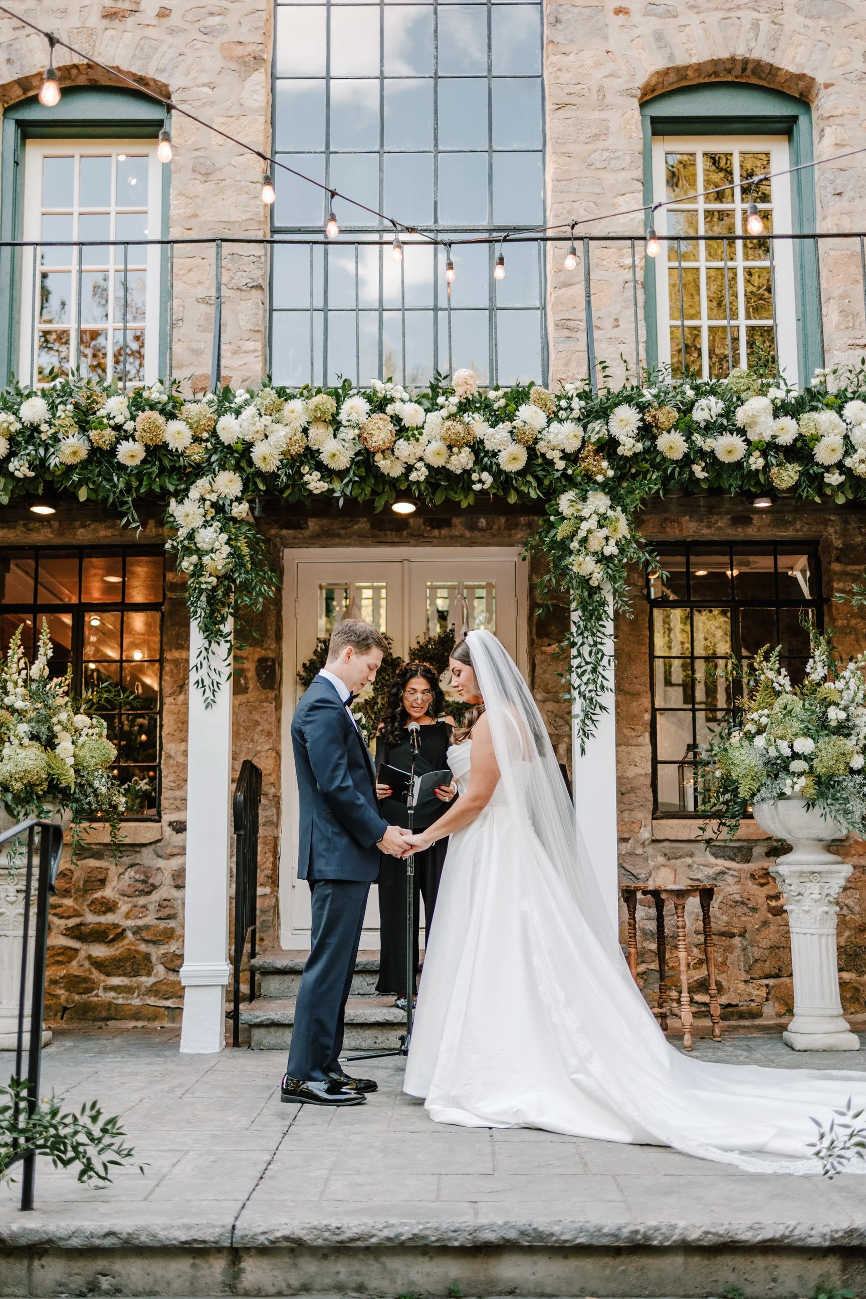 A bride and groom holding hands during a wedding ceremony outdoors, with an officiant standing behind them. The setting includes floral decorations and a stone building with large windows.