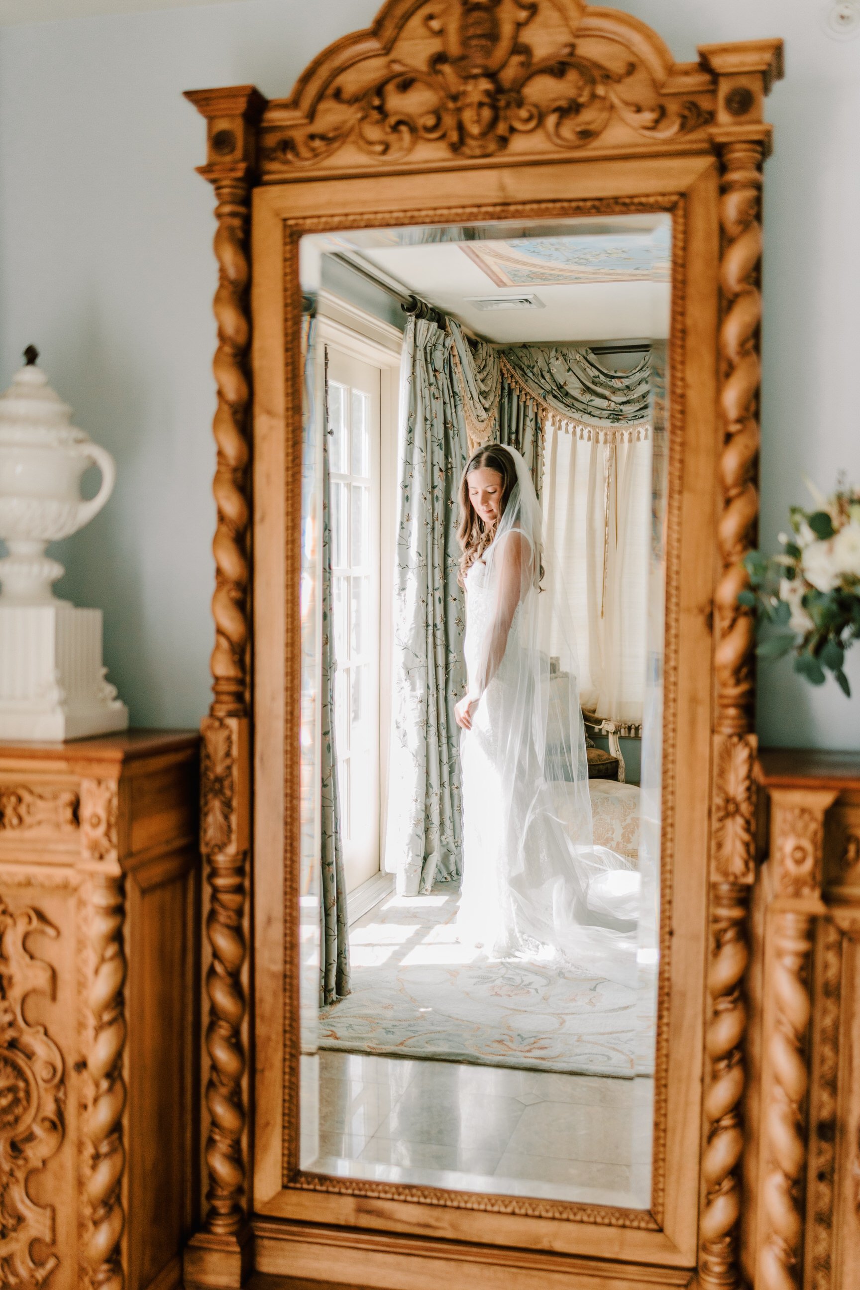 A bride in a white wedding dress and veil stands in a well-lit room, looking down with her eyes closed, seen through a large, ornately carved wooden mirror.