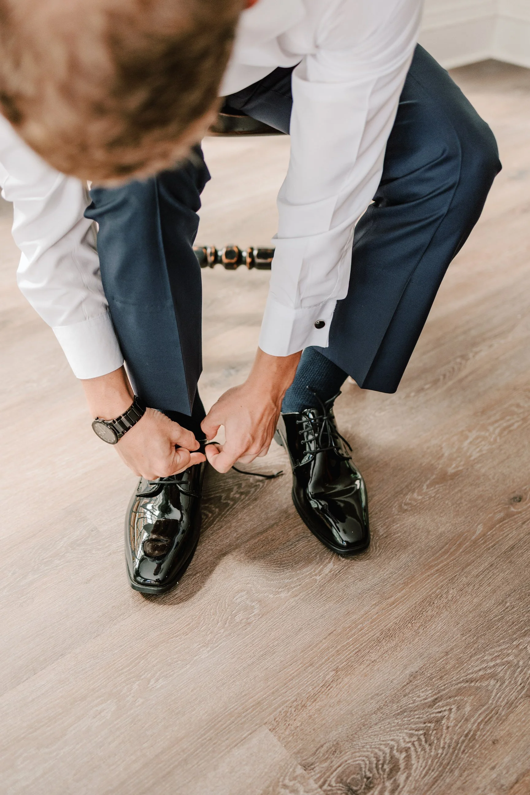 Man in formal attire tying his shiny black dress shoes, with dark dress pants, white shirt, and watch, on a wooden floor.