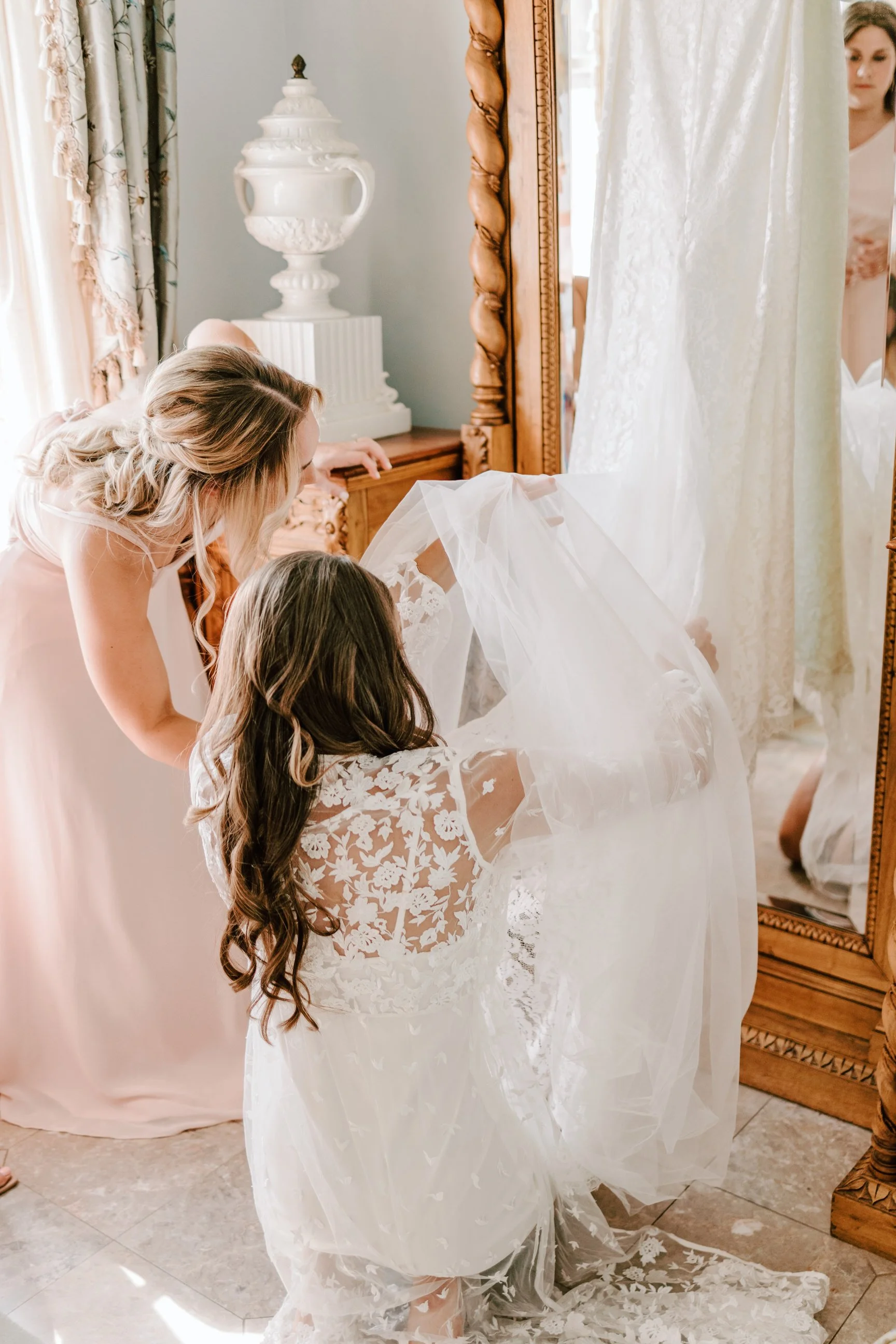 A bride in a lace wedding dress kneeling while another woman helps her with her veil in a bedroom with a large mirror and a wooden dresser.