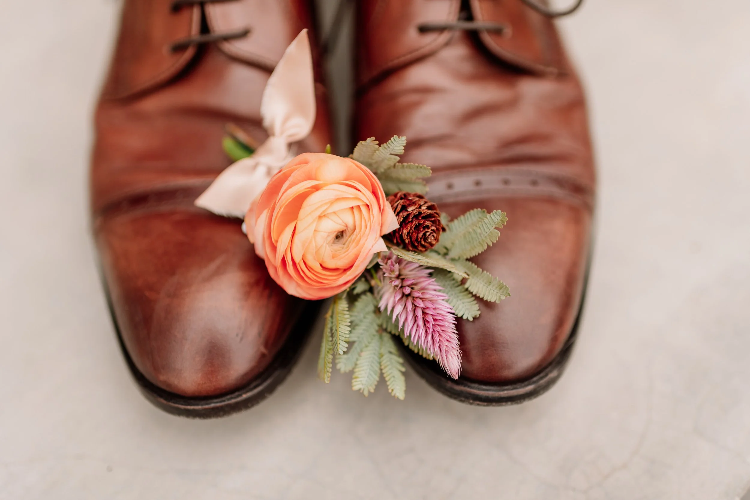 Pair of brown leather dress shoes with pink and orange floral boutonniere on the toe, including a peach ranunculus, pink astilbe, pine cone, and fern leaves.