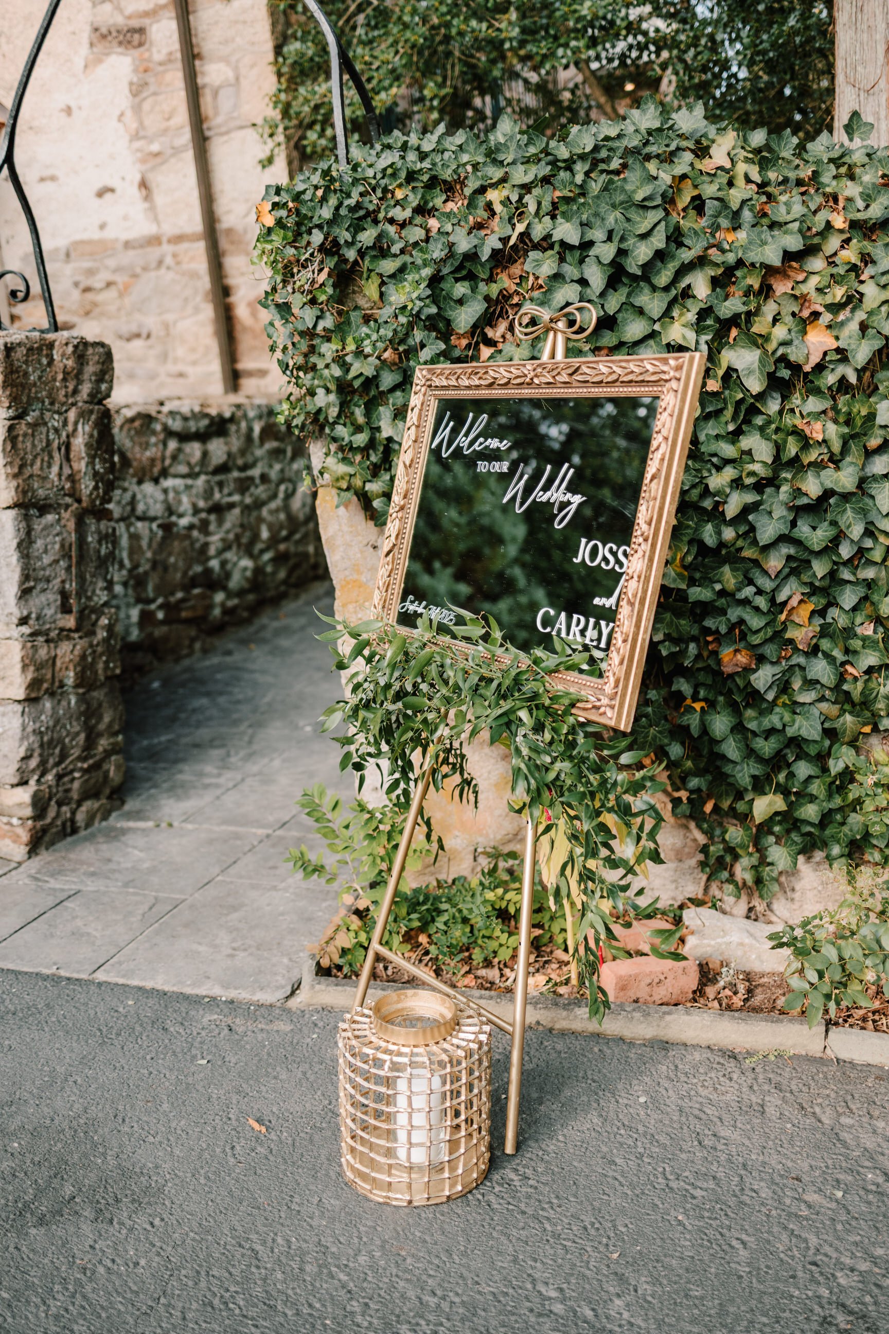 Wedding welcome sign with greenery and a candle in a decorative holder at an outdoor venue.