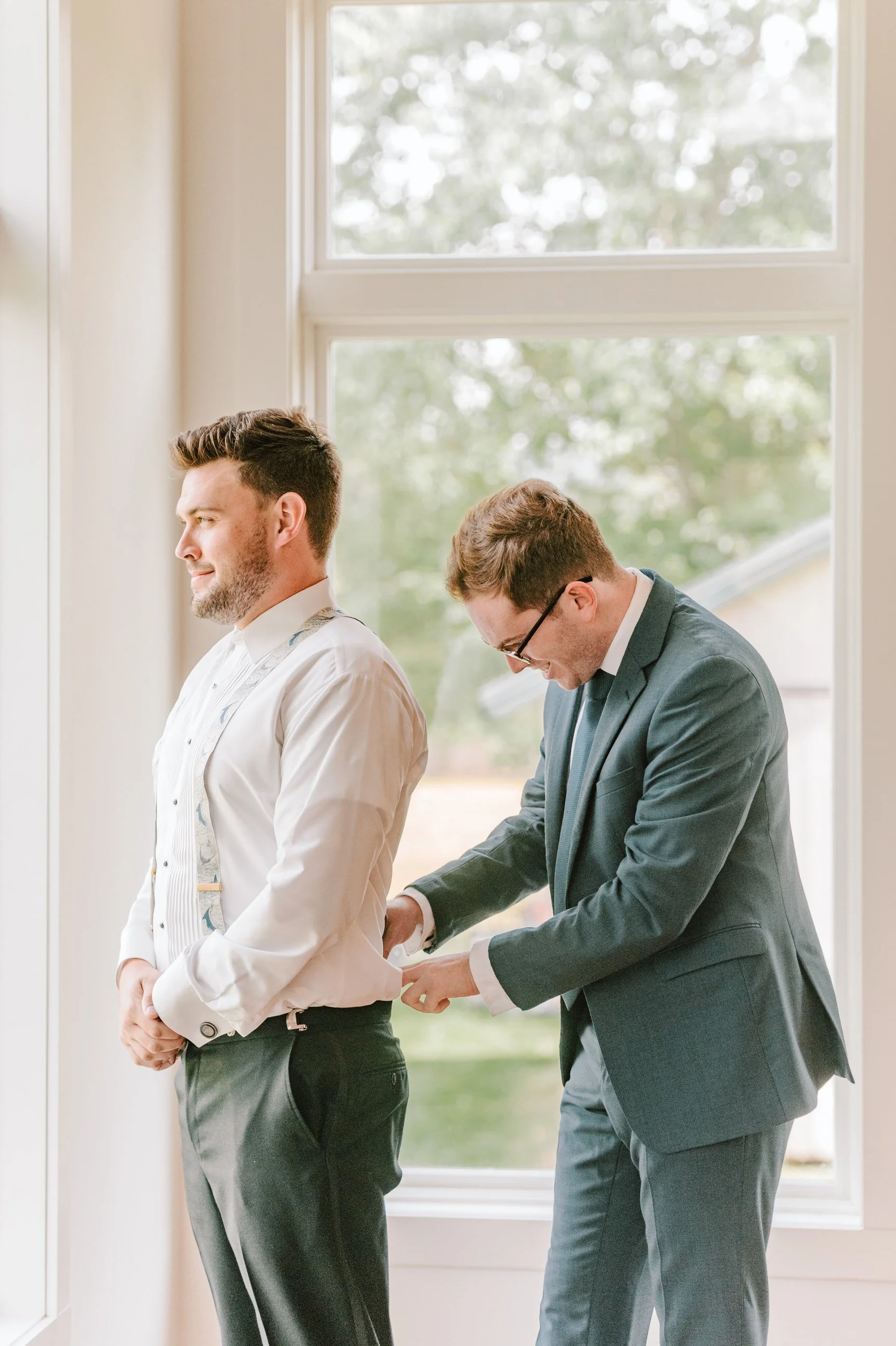 Two men, one helping the other with cufflinks by a large window, indoors. One man is dressed in a white shirt and the other in a dark suit.