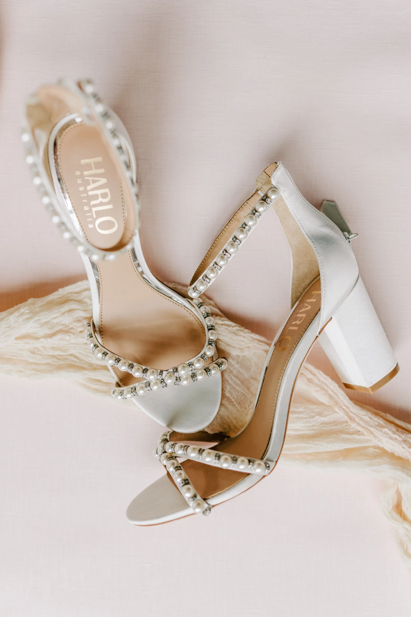 A pair of white high-heeled shoes decorated with pearl and rhinestone embellishments, placed on a textured fabric surface.