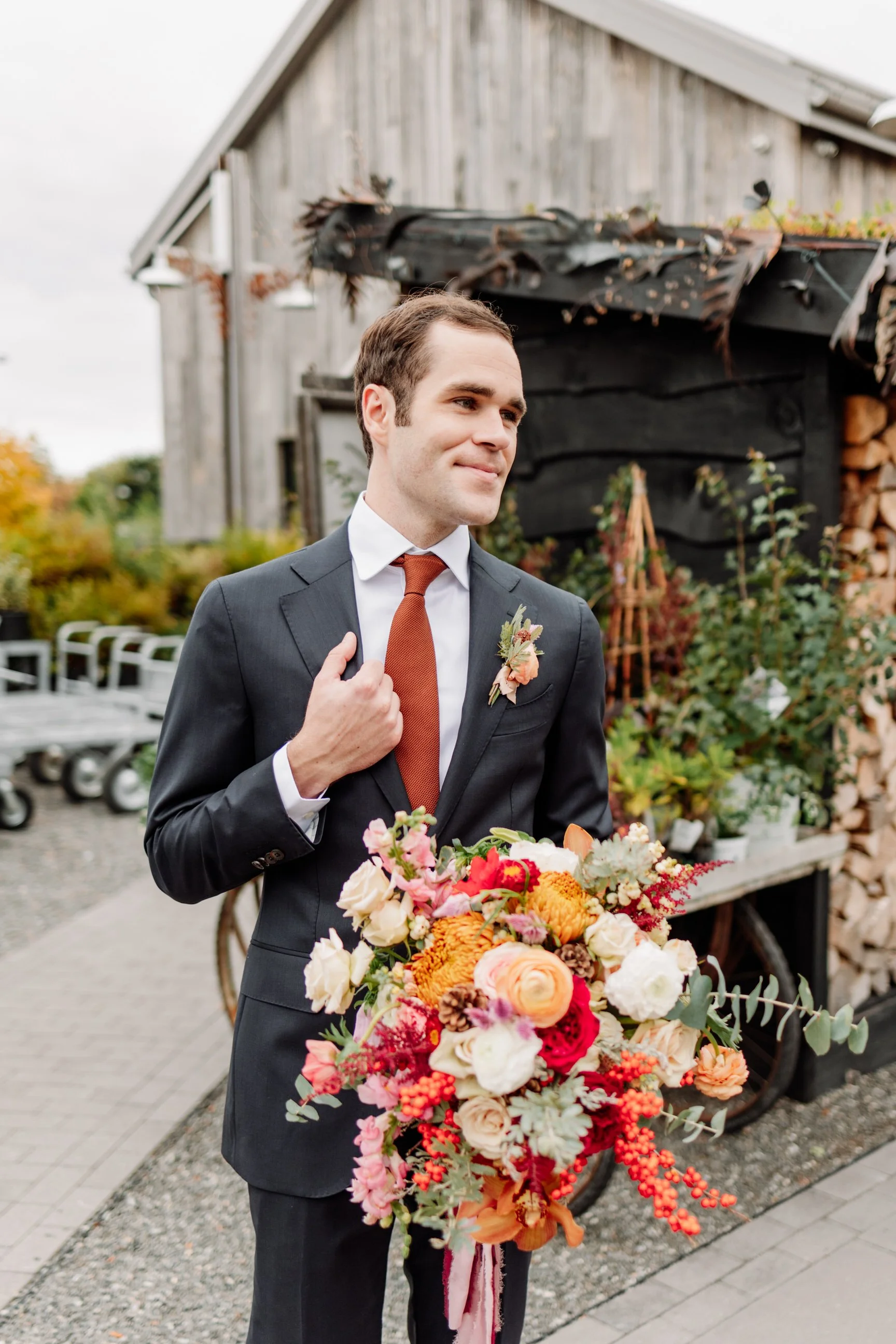 A groom in a dark suit holding a large bouquet of colorful flowers in front of a rustic wooden building.