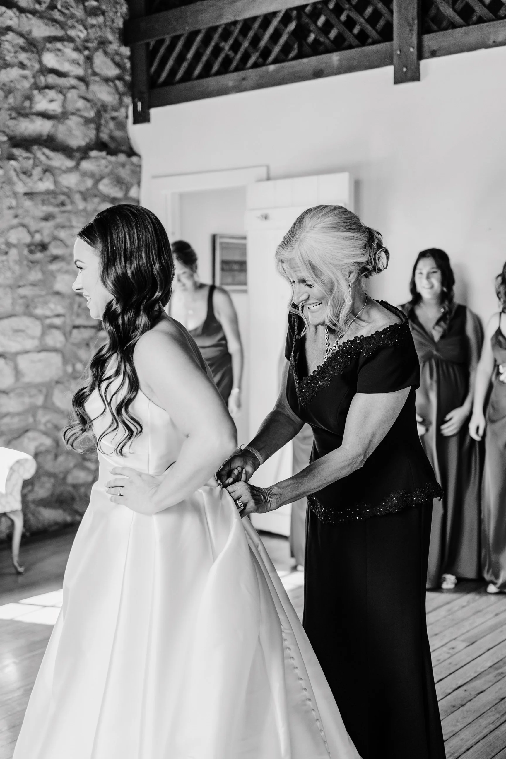 A bride in a wedding dress standing with her hands on her hips as an older woman helps adjust her dress, while three women in dresses look on and smile in a rustic indoor setting.