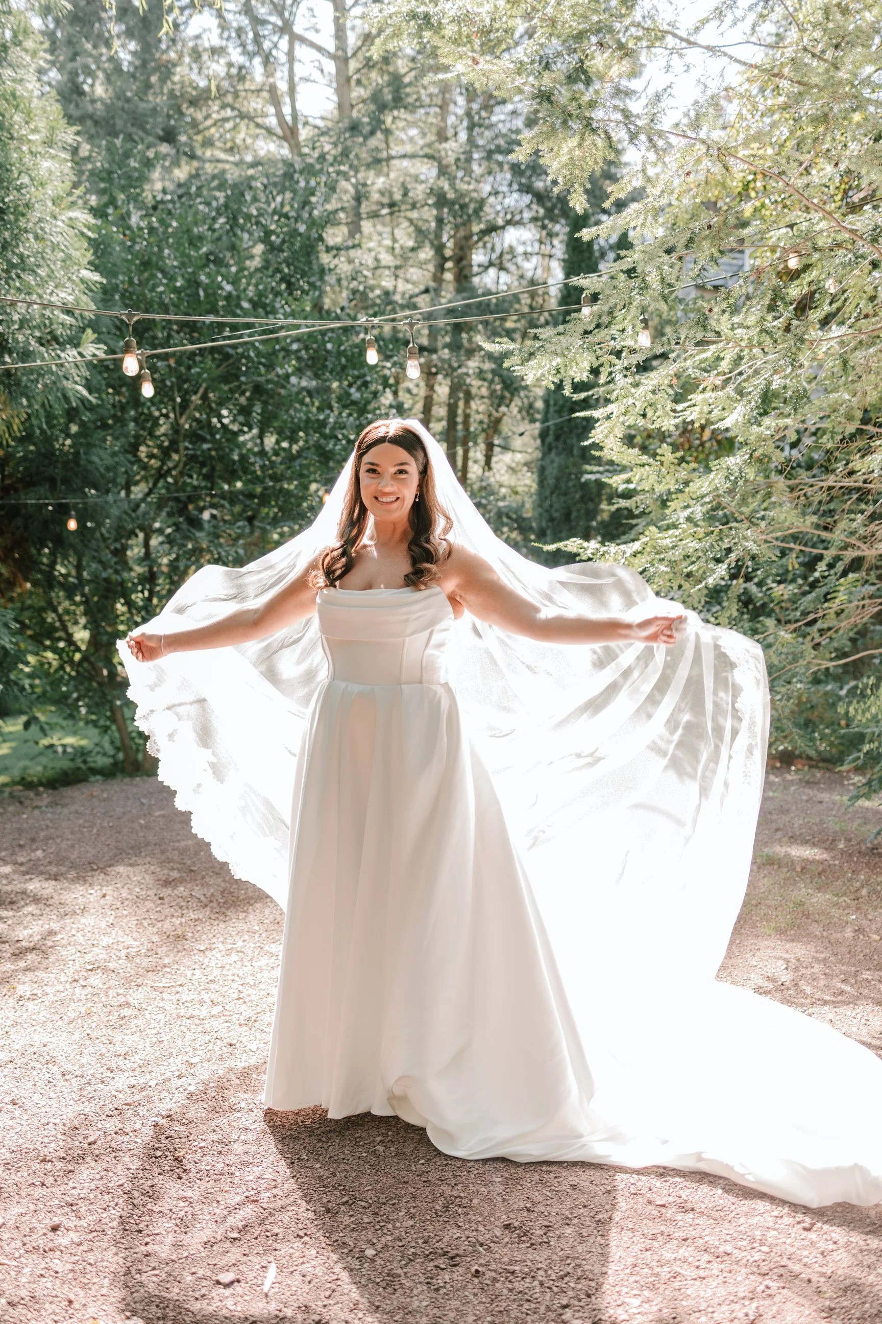 A smiling bride in a white wedding gown holding her veil in an outdoor garden setting with string lights and green trees in the background.