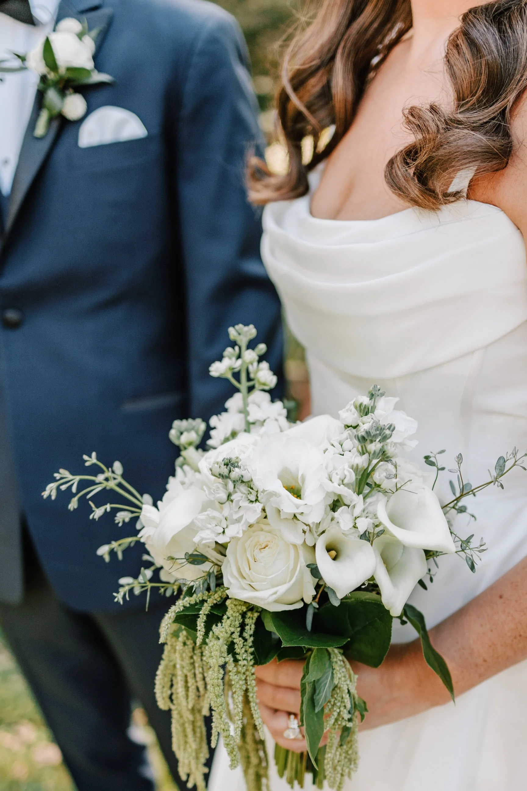 Bride holding a bouquet of white roses, calla lilies, and greenery, with groom in a navy tuxedo standing nearby.