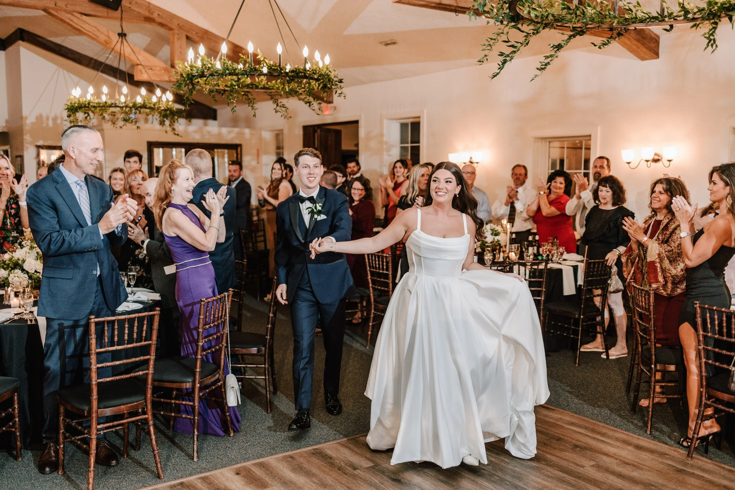 A bride in a wedding dress is holding hands with a groom in a tuxedo, walking into a reception with family and friends applauding and smiling.